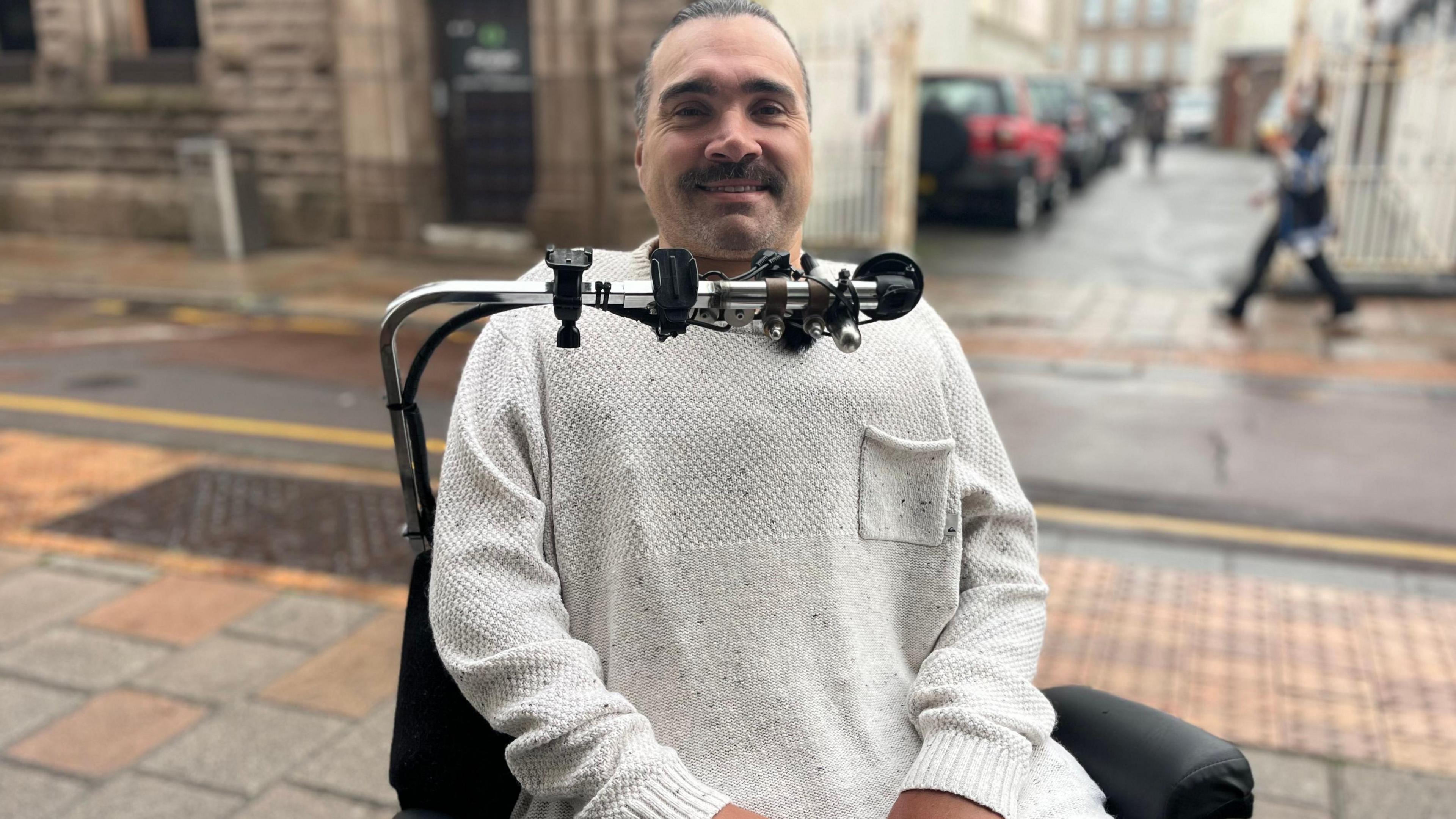 James Bedding is a man with dark eyebrows and grey hair. He is wearing a cream jumper. He is sitting in a wheelchair with a pole in front of his face that has a stick he can use to steer his wheelchair with. The background is an out-of-focus town street.