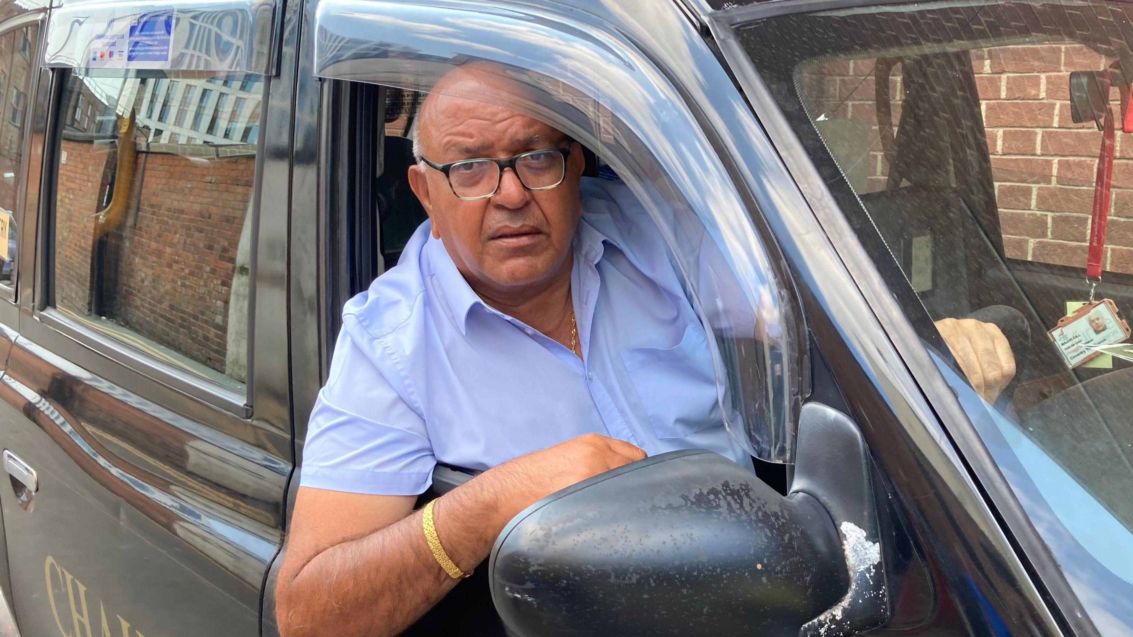 An Asian man wearing glasses and with bald head and grey hair on the sides, is wearing a pale blue short leave shirt. He is leaning his right elbow out of the drivers side window of a black cab window.