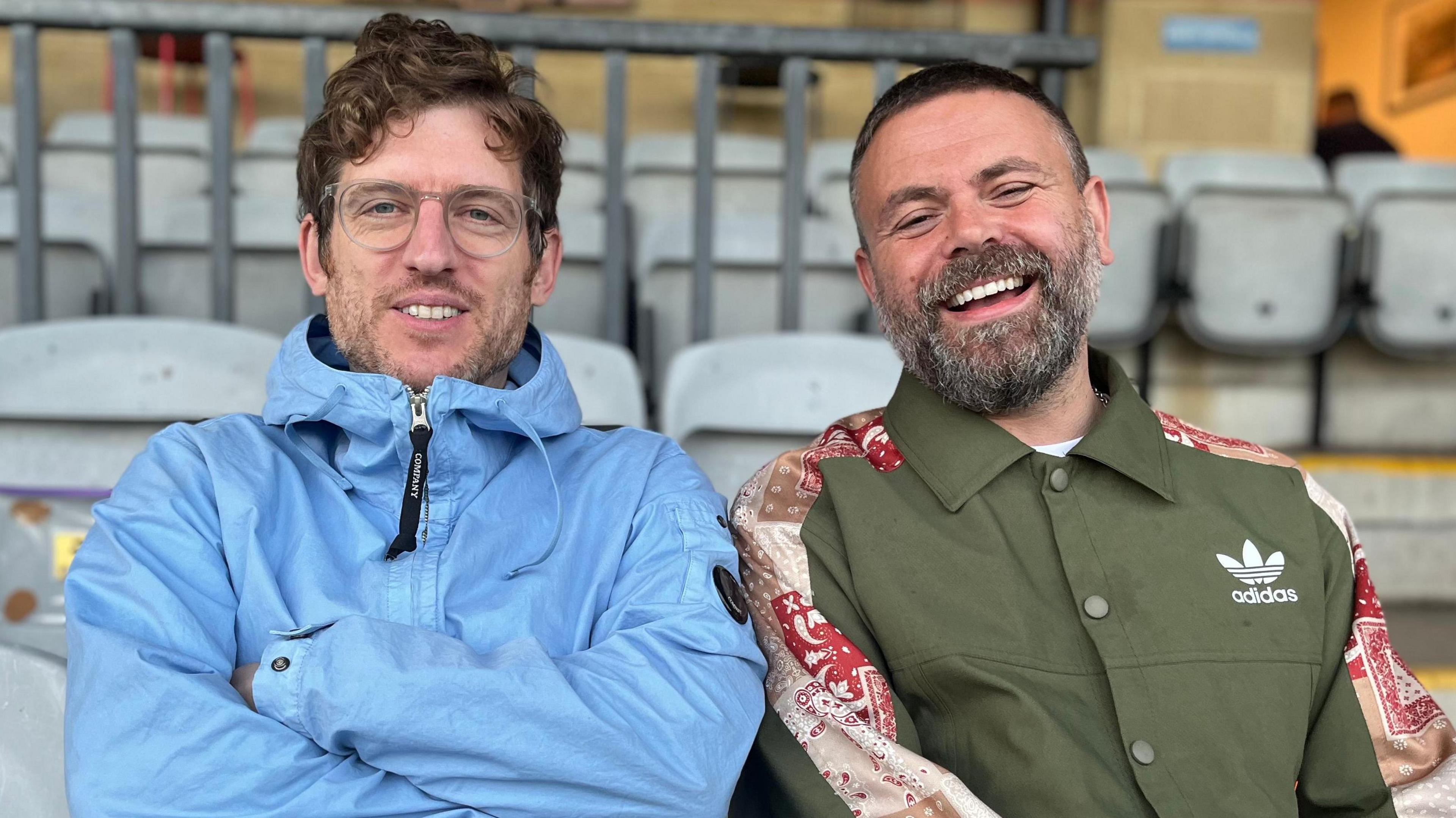 Geraint John Elis James are sitting in football stands, looking ointo the camera and laughing. Geraint has short dark greying hair and a beard. He is wearing a green buttoned-up jacket with red detailing on the shoulder. Elis is wearing glasses with a translucent rim and and light blue waterproof jacket with a hood. He has brown curly hair.