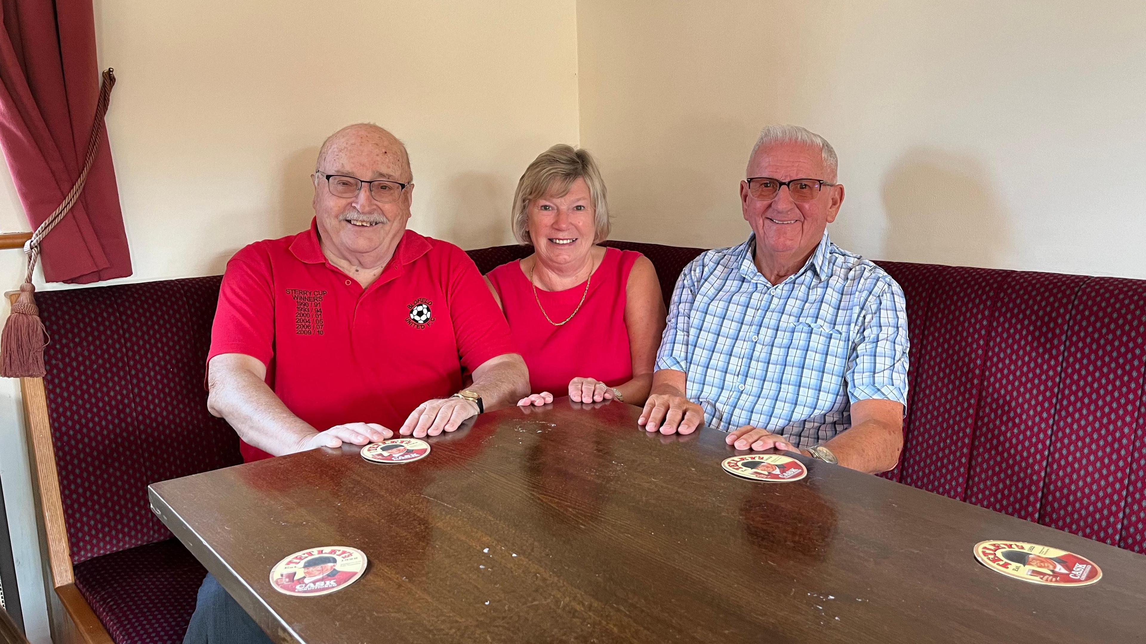 Blofield FC president Paul Roofe wearing a red club polo shirt, Paddy's wife Jenny Murphy, also wearing a red top, and Acle Rangers chairman Colin Tovell, in a blue-checked shirt, sit together on a bench in Blofield FC Club House. 