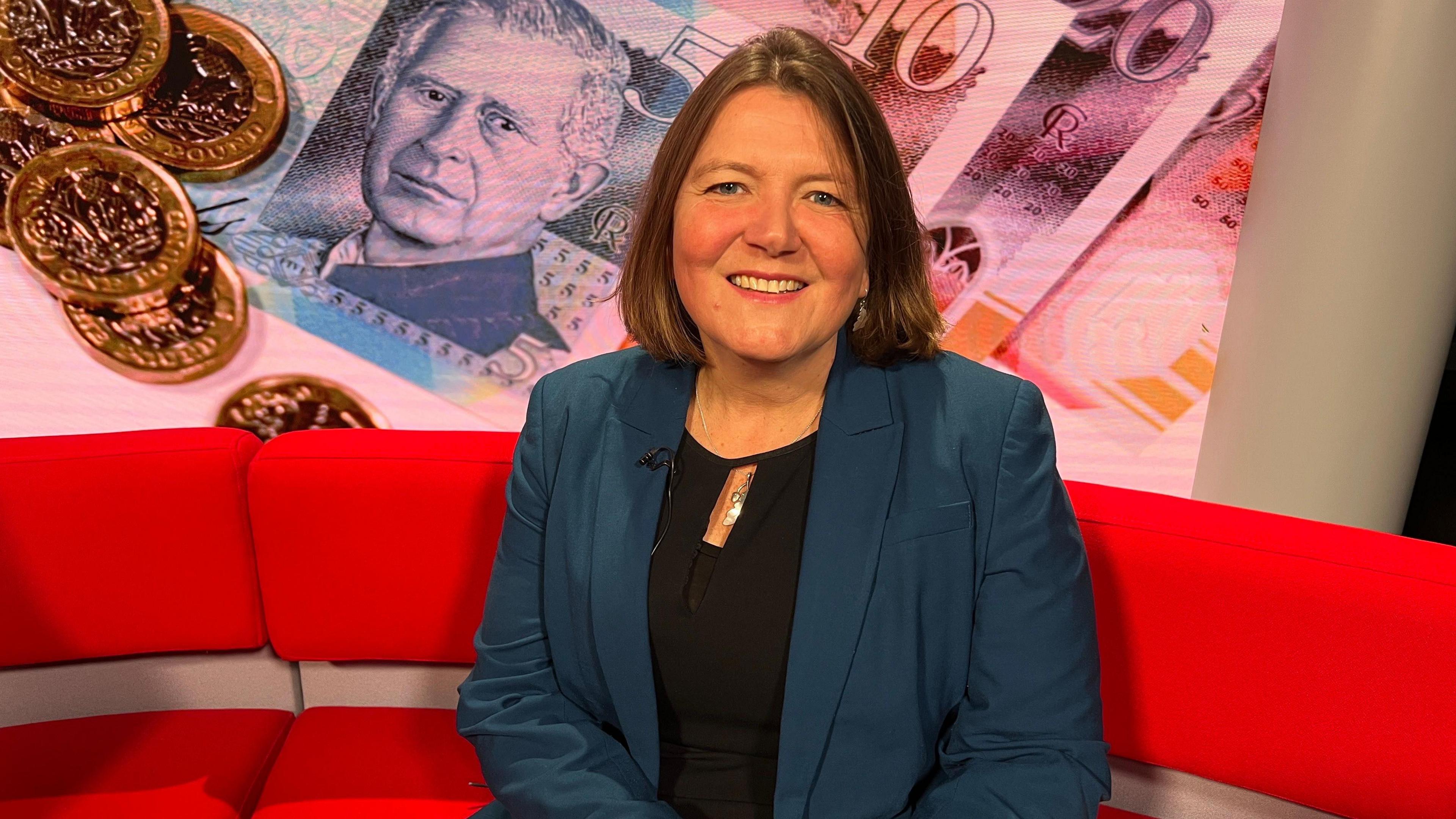 A woman with short light brown hair. She is sitting on a red sofa and is wearing a black blouse with a dark teal suit jacket