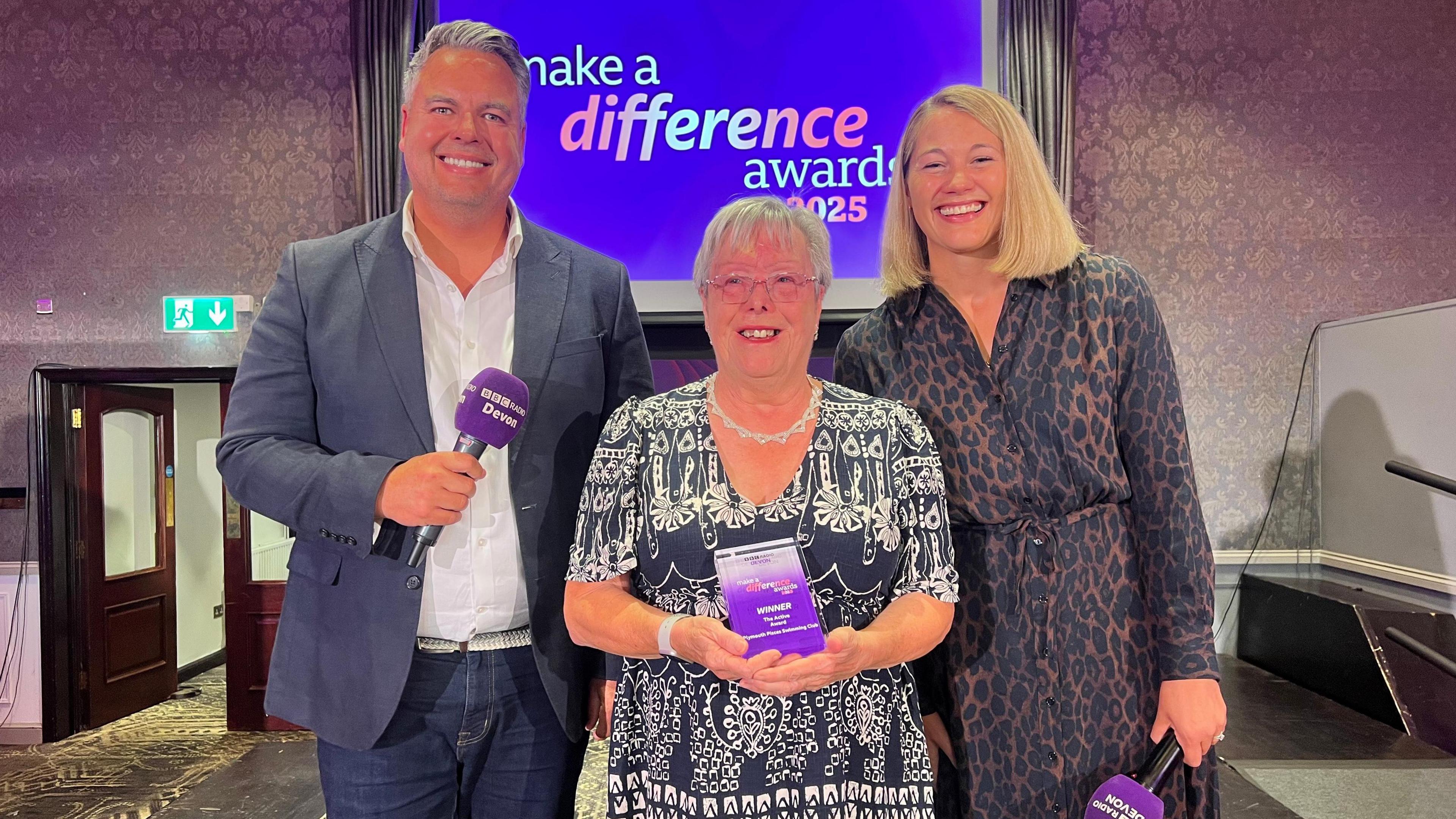 Two women and a man stand together on stage at the make a difference awards. The woman in the middle is smiling through tears. She is holding a rectangular trophy.