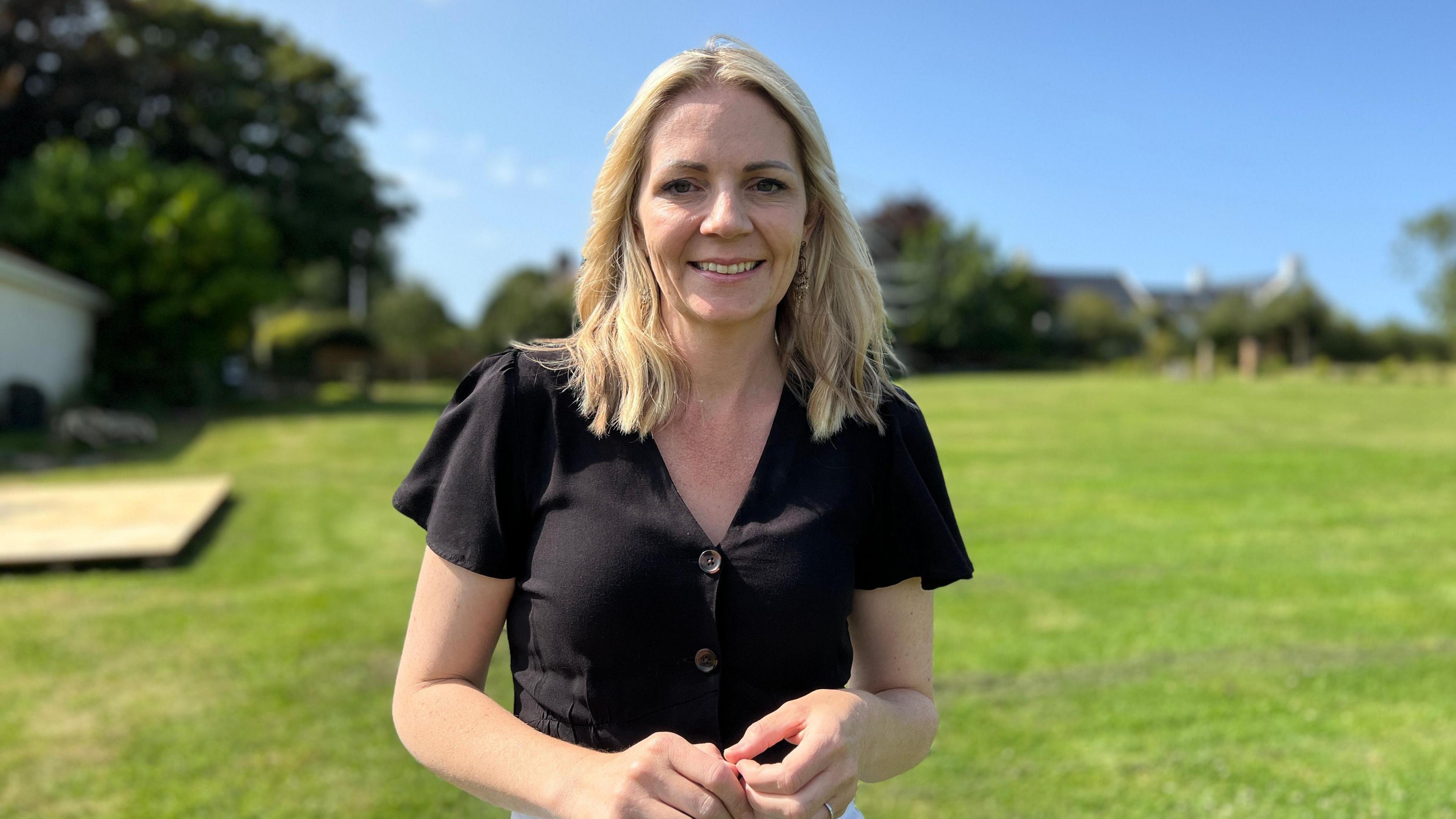 Chloe smiles at the camera with a field in the background. She is wearing a black shirt and she's holding her hands. Her blonde hair is being blown slightly in the wind.