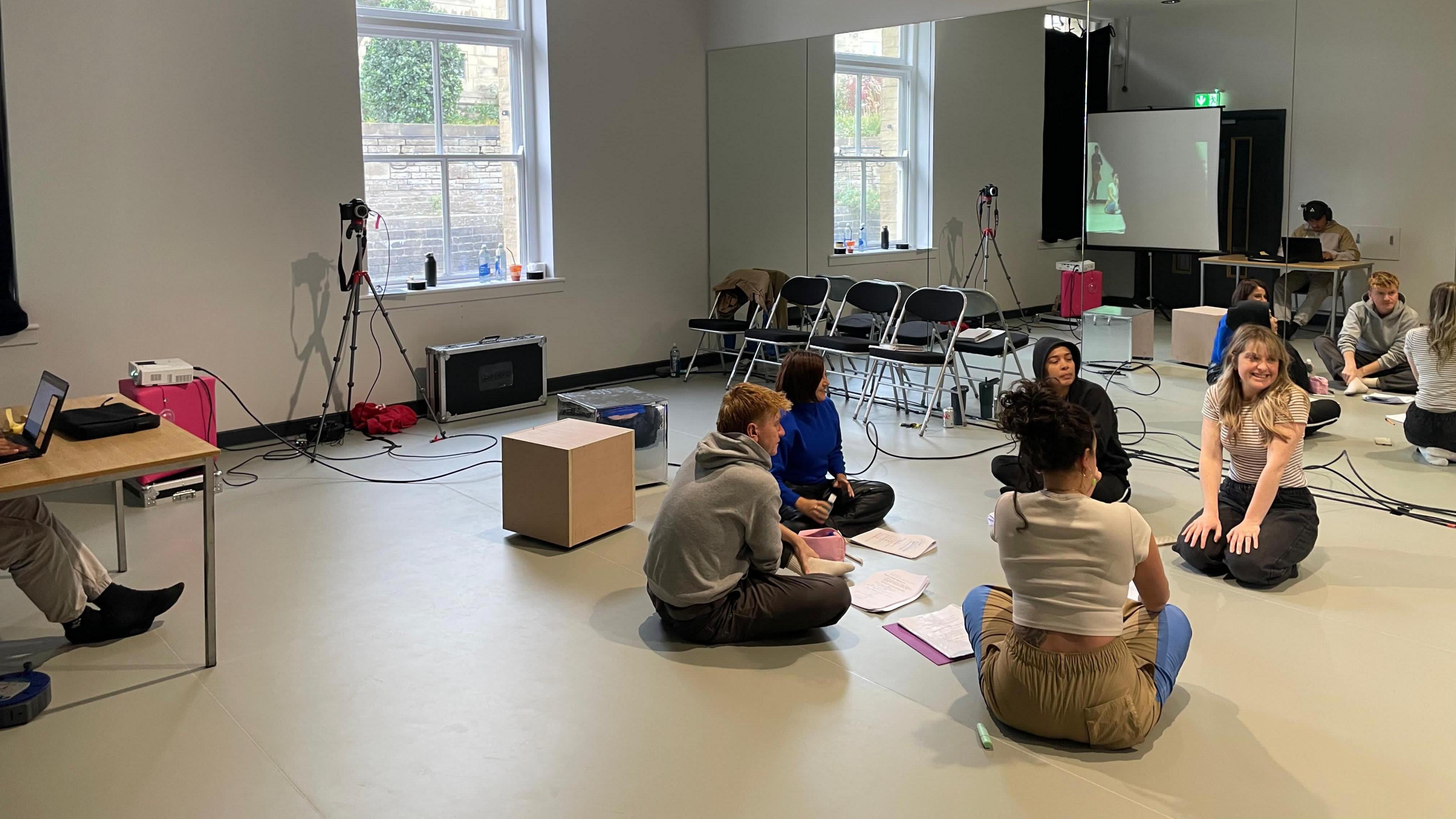Five people are sitting on the floor in a rehearsal studio. There is a mirror across the wall behind them and four chairs in the corner.