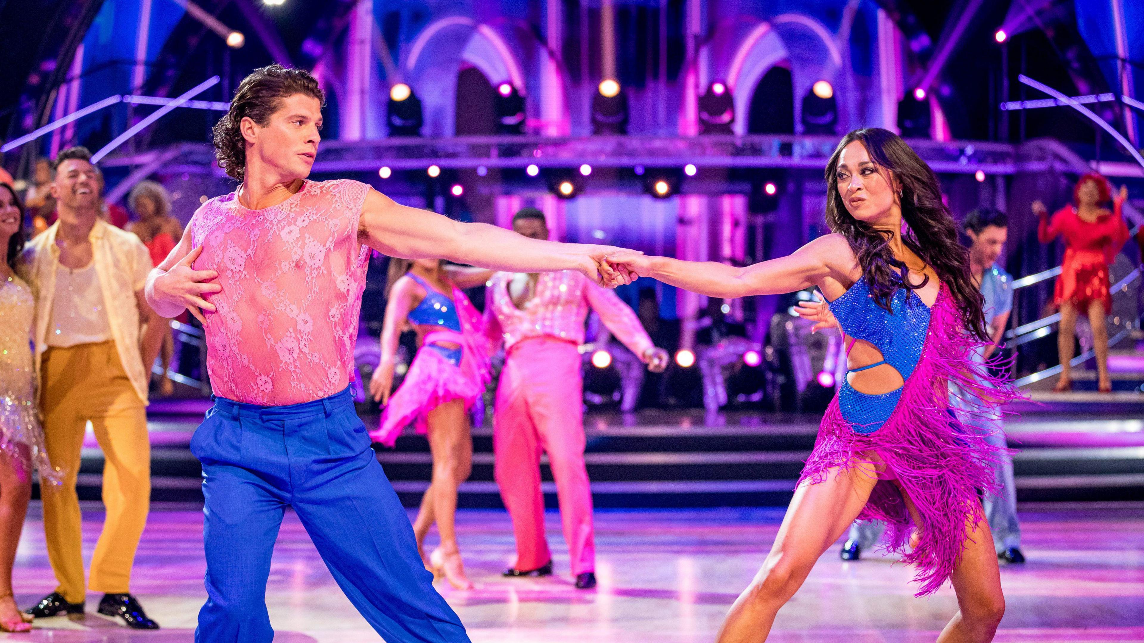 Lewis Cope & Katya Jones on the launch show of the new series of Strictly Come Dancing - the two are in the middle of the group dance, both wearing a dazzling combination of pink and blue, holding hand with their arms extended to maximum length, looking at each other in the middle of a dance move