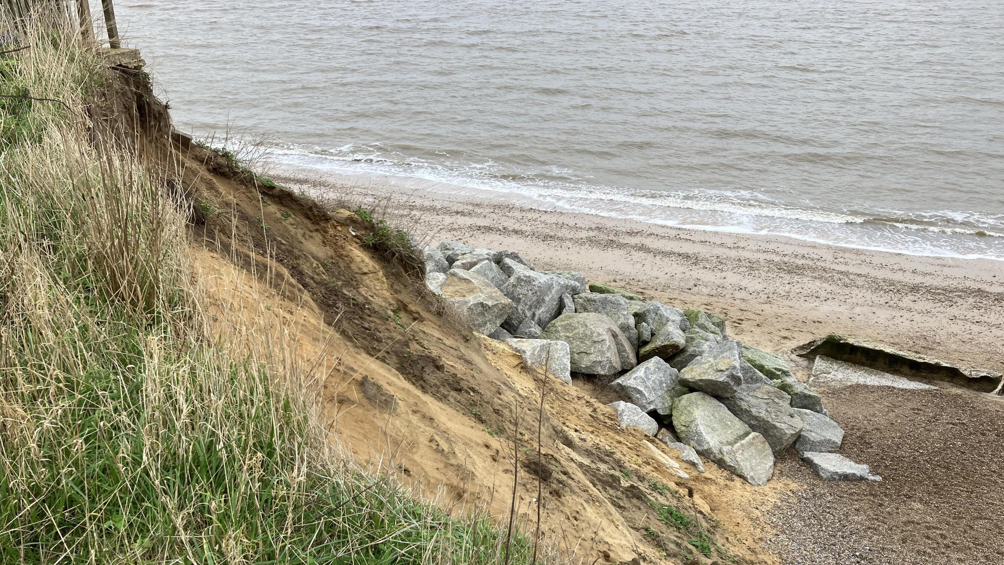 An eroding, sandy cliff with a pile of grey, granite rocks at the bottom, with a gravel beach and sea in the background 