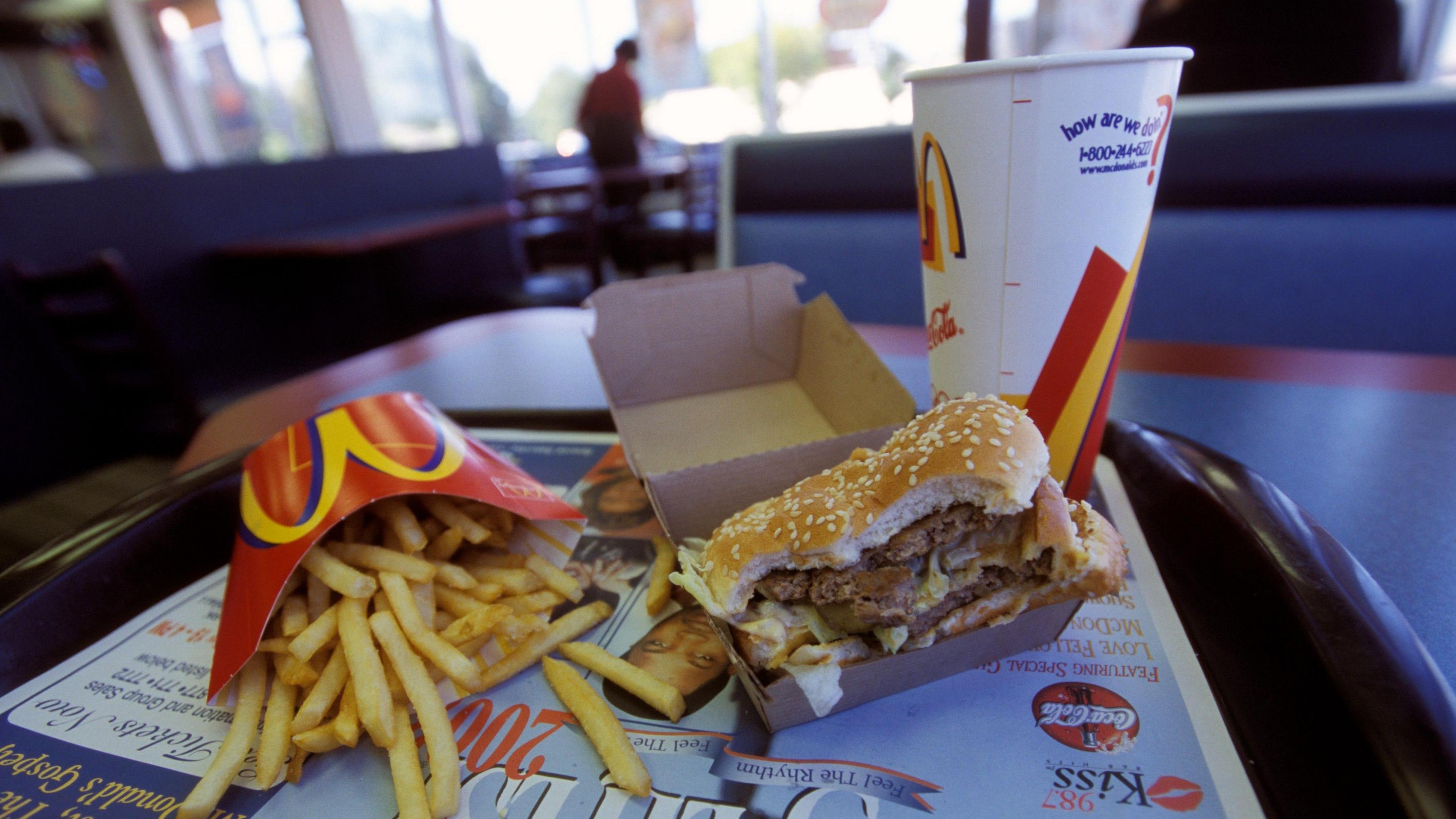 A McDonald's meal sits on a tray in a fast food restaurant, the burger is half eaten, the fries are spilling out of the cardboard folder and a soda drink stands behind both.