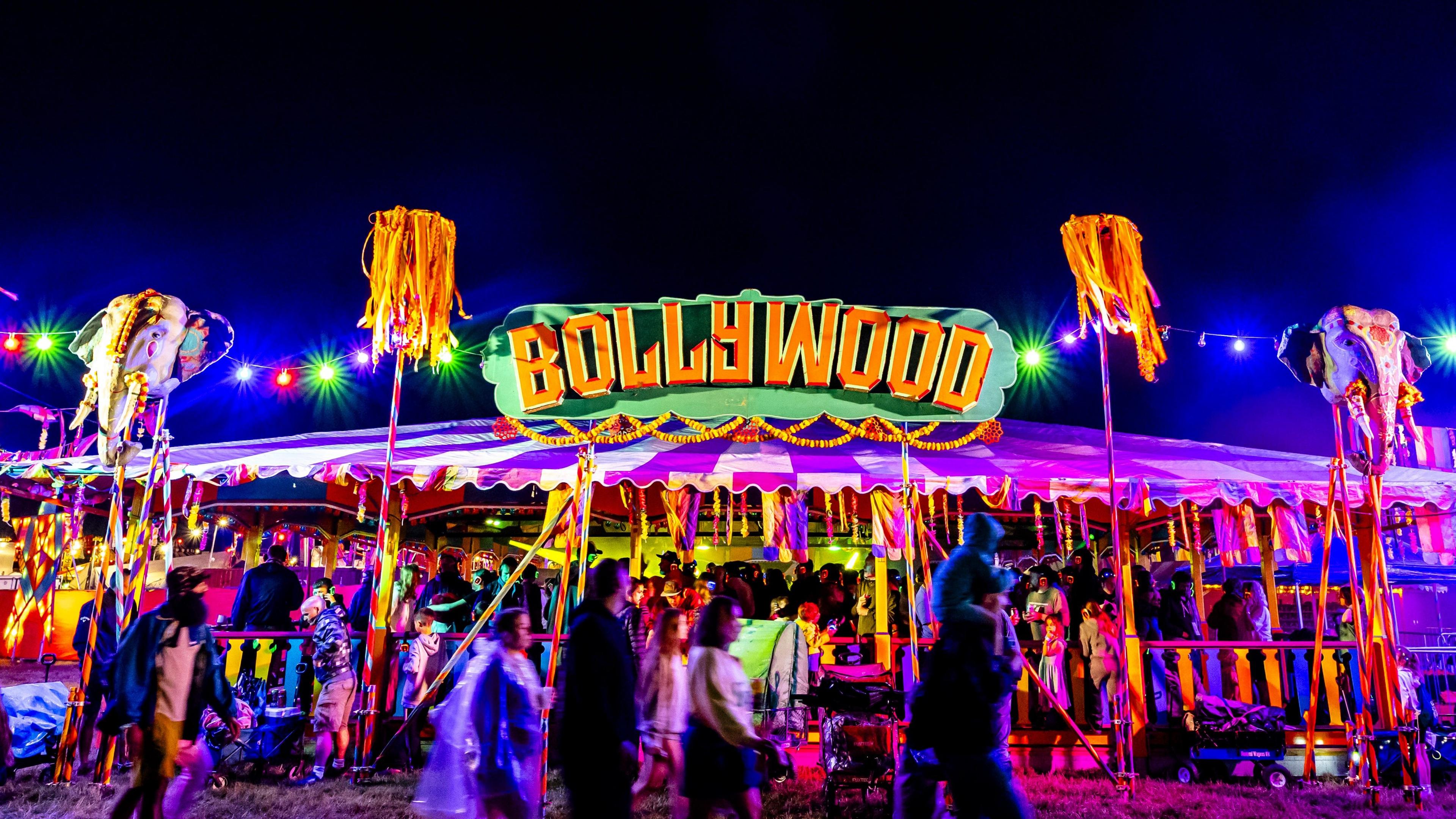 A circus-style tent of red and white is reflected with neon pink light. On the top of the tent is a sign reading "Bollywood" in green and yellow light. There are colourful tassels hanging down from the tent, and inside, there are hundreds of people