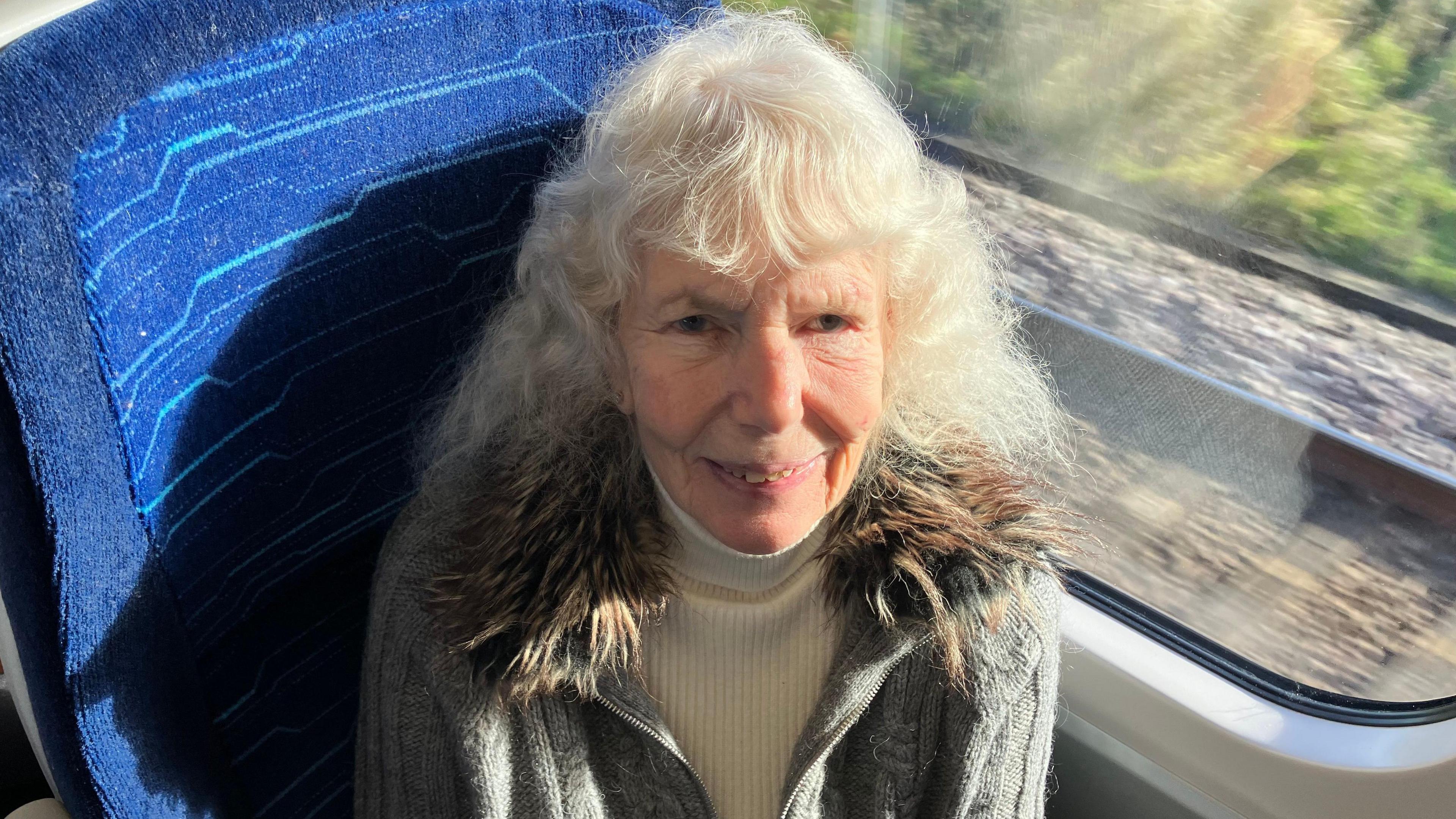 Big Communi-Tea client Ann Waddington on the train to London. She is sitting in a window seat, smiling, and is wearing a white polo neck and a grey cardigan with a fur-trimmed collar.