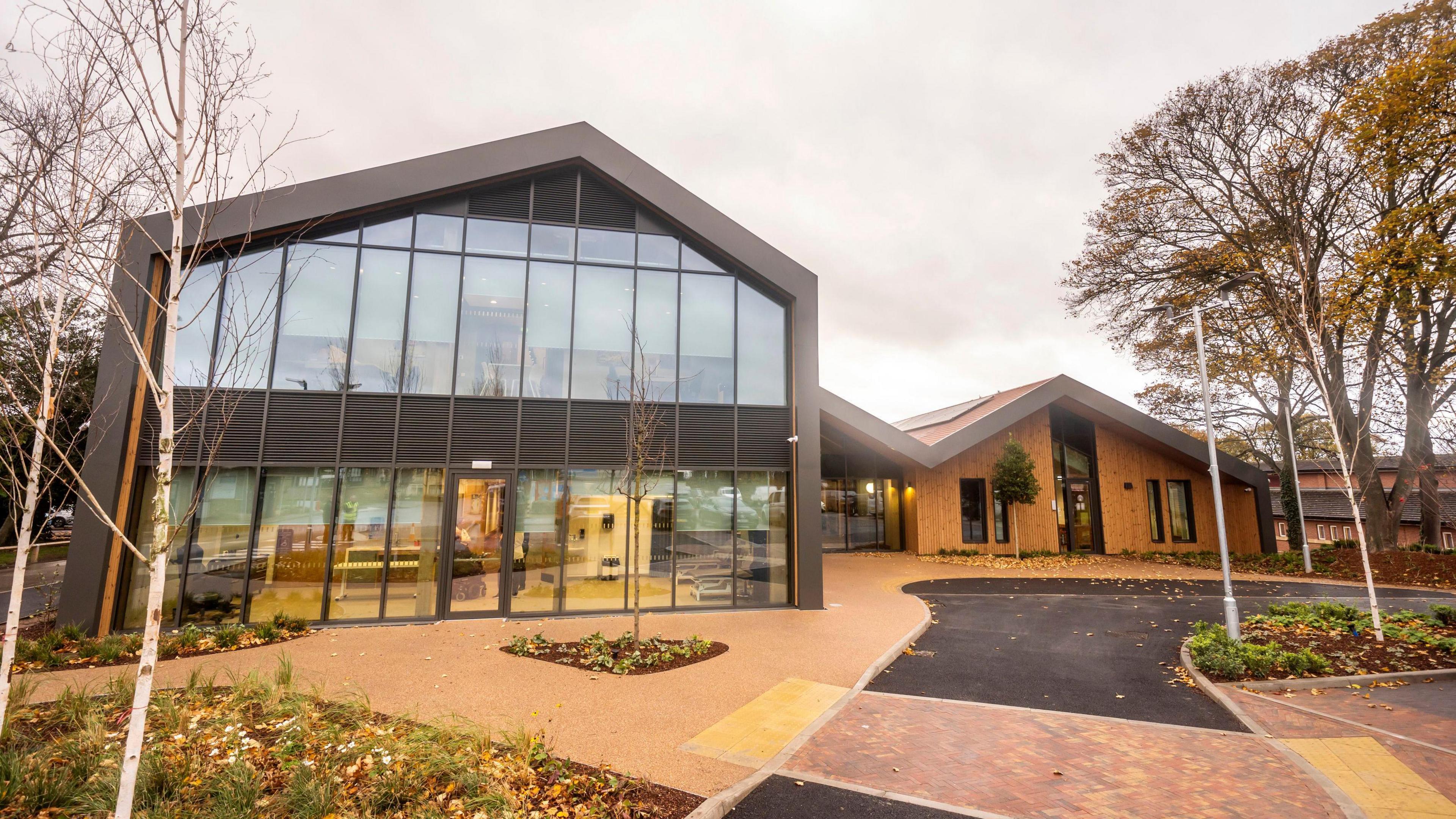 The new Rob Burrow MND Centre, pictured from the outside. A large glass building is prominent in the picture, with a smaller shed-like building further behind. Footpaths and plants can also be seen in the foreground.