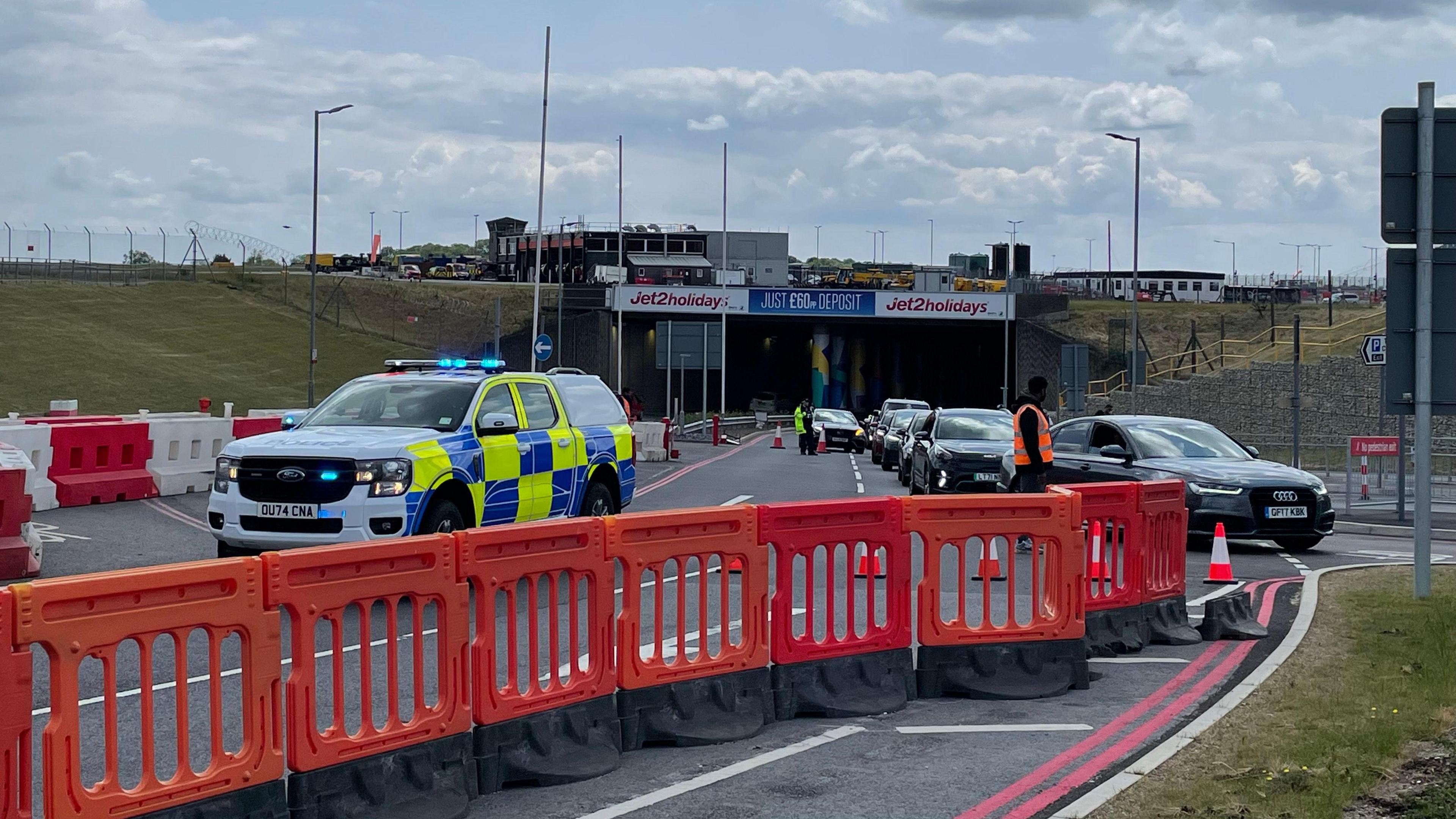 A police truck has its blue lights on behind a line of orange barriers shutting the road. It is blocking the entrance to a tunnel at the airport, which has a stream of cars queuing to get out.