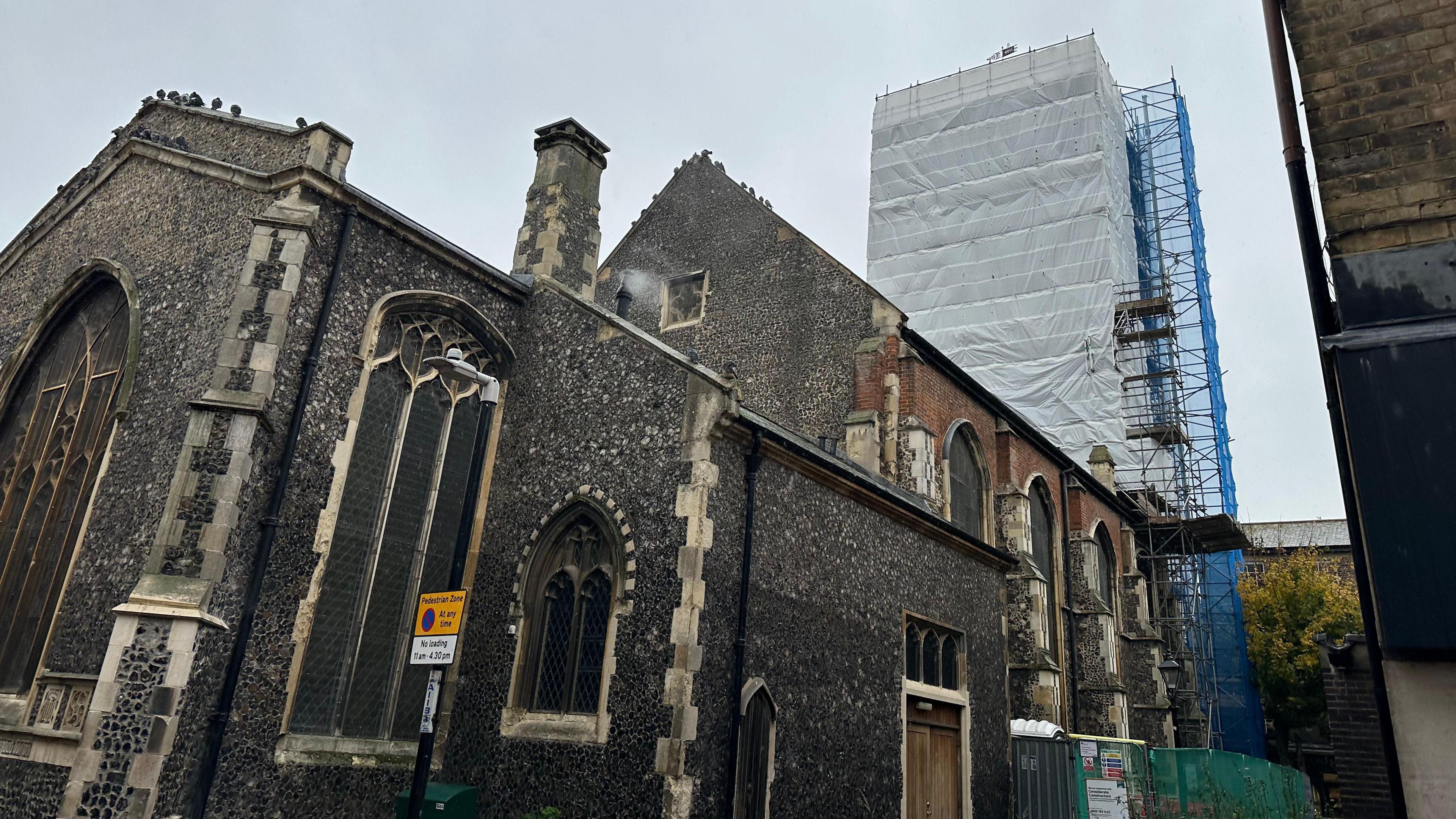 A black stone medieval church building with a tower in the background - scaffolded in white and blue.