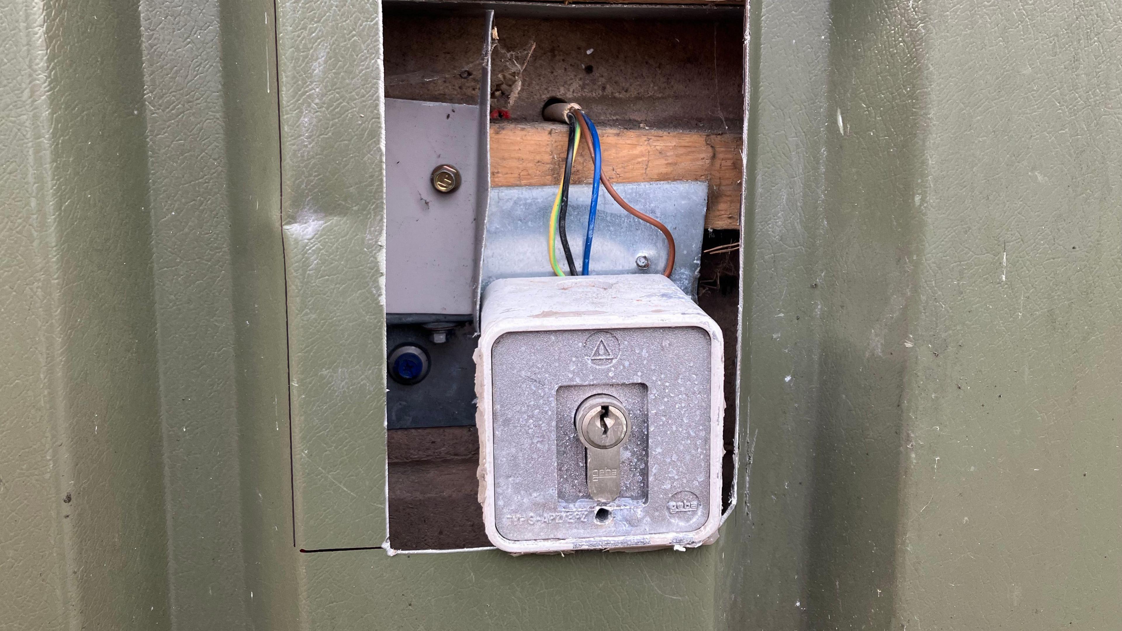 A rectangular hole has been cut in a green metal casing exposing a lock attached to yellow, black, blue and brown wires.