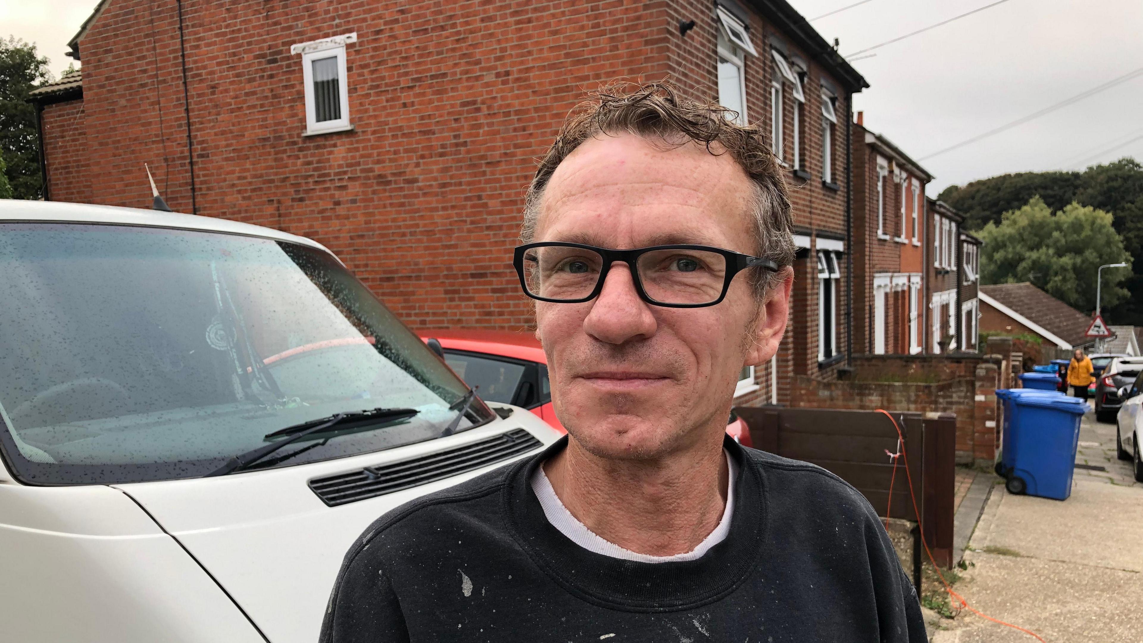 A general view of Kevin Hollock. He is facing the camera and smiling. Behind him is a driveway with a grey van and a red car parked. Houses line the street behind him while another person walks toward him on the pavement. He is wearing black glasses as well as a black jumper with a white t-shirt underneath. He has brown and grey short curly hair. 