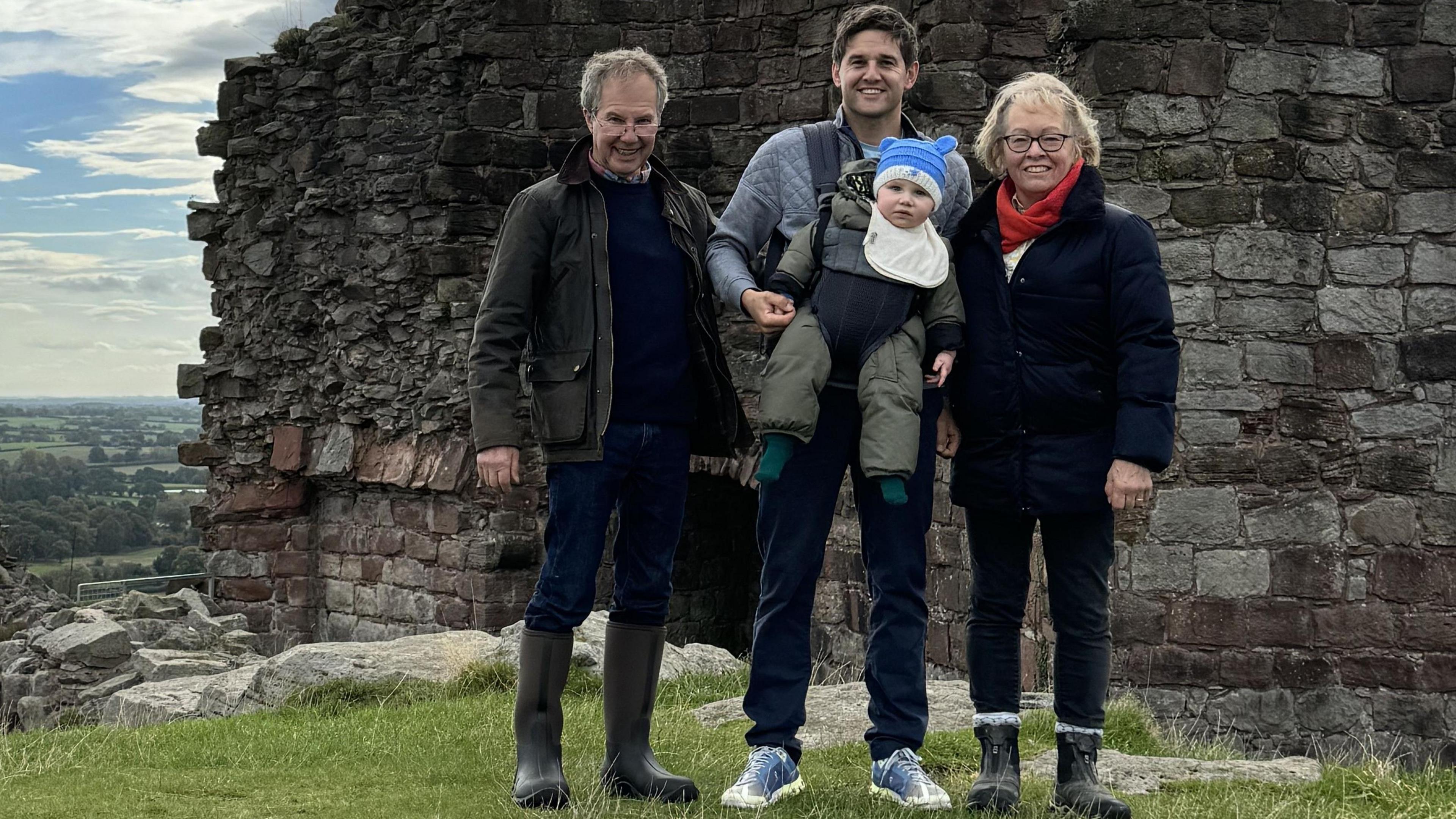 A family group standing outdoors in front of the remains of an old stone wall, part of an historic ruin or castle. The people are dressed in outdoor clothing, including jackets and boots, and the man in the centre is carrying a small child. Behind the group there is a scenic view of green fields stretching into the distance under a partly cloudy sky. 