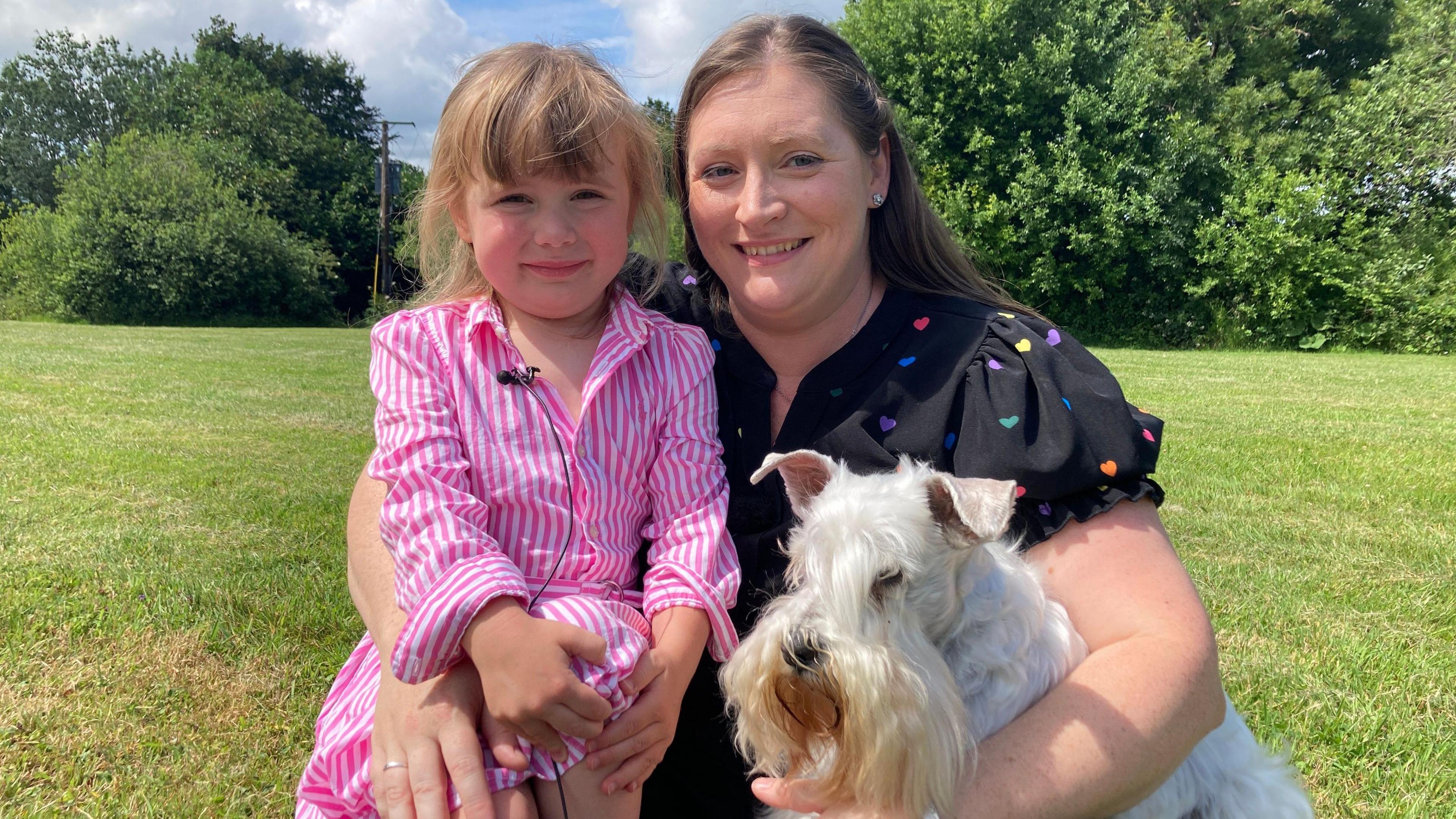 Caitlyn in a pink and white striped dress is sat cuddling a woman with long hair who is wearing a black top with coloured hearts on it. The woman has her other hand around a white dog. Grass and trees can be seen behind them.