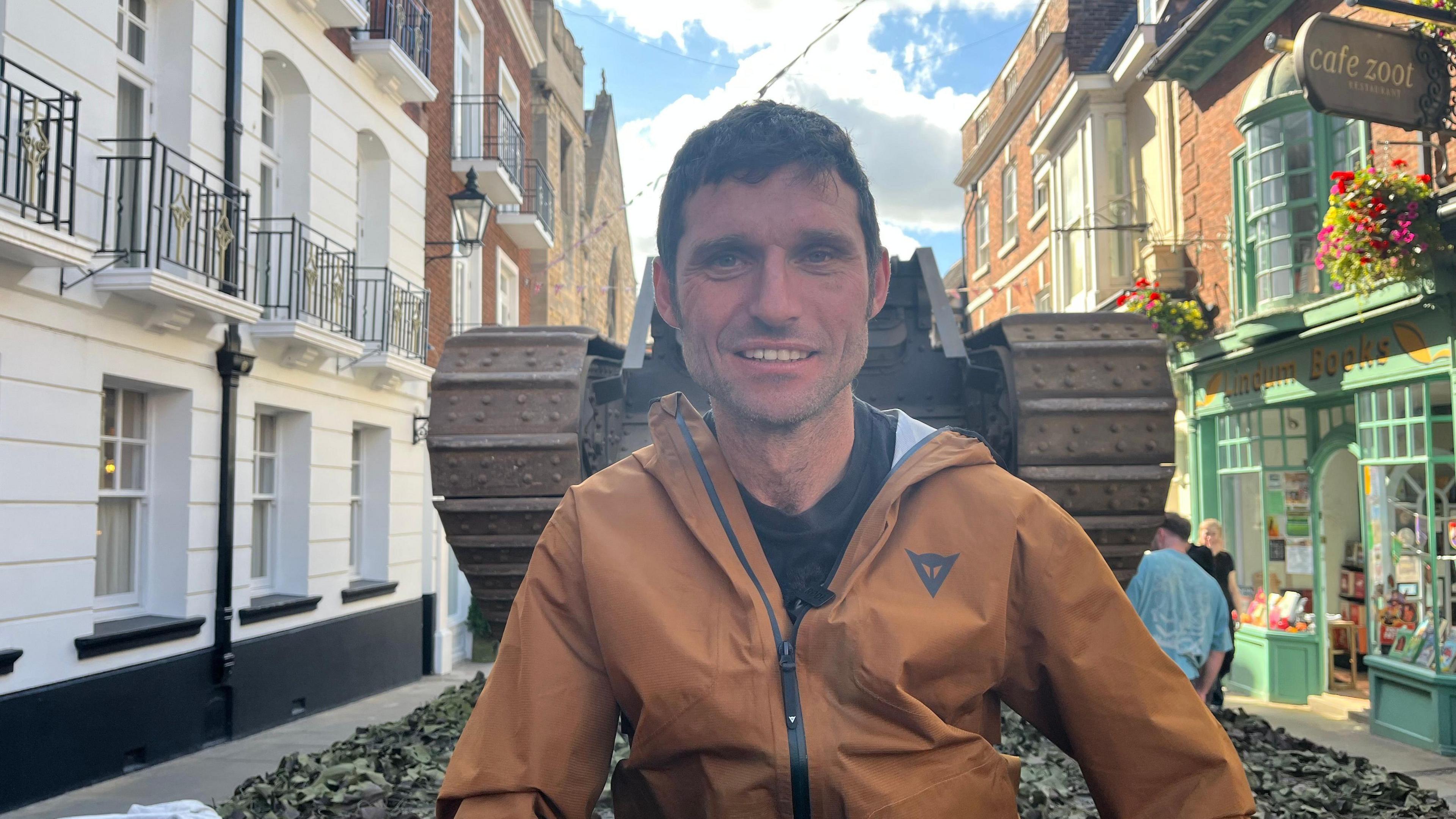 A man with short dark hair wears a black t-shirt and bronze coloured jacket. He is sitting on the back of lorry which is carrying a vintage tank. The vehicle is travelling through a town centre street lined with historic buildings.