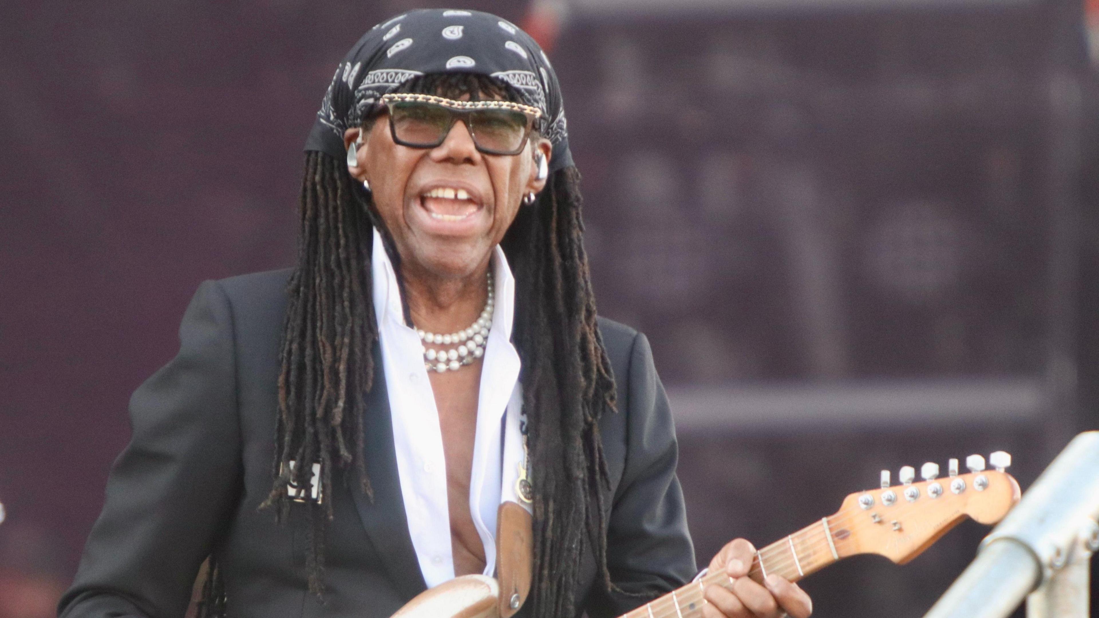 Nile Rodgers performing with is guitar, singing and looking into the camera. He has a black and white bandana over his hair, sunglasses and a white necklace on. He is wearing an open top white shirt and black blazer.