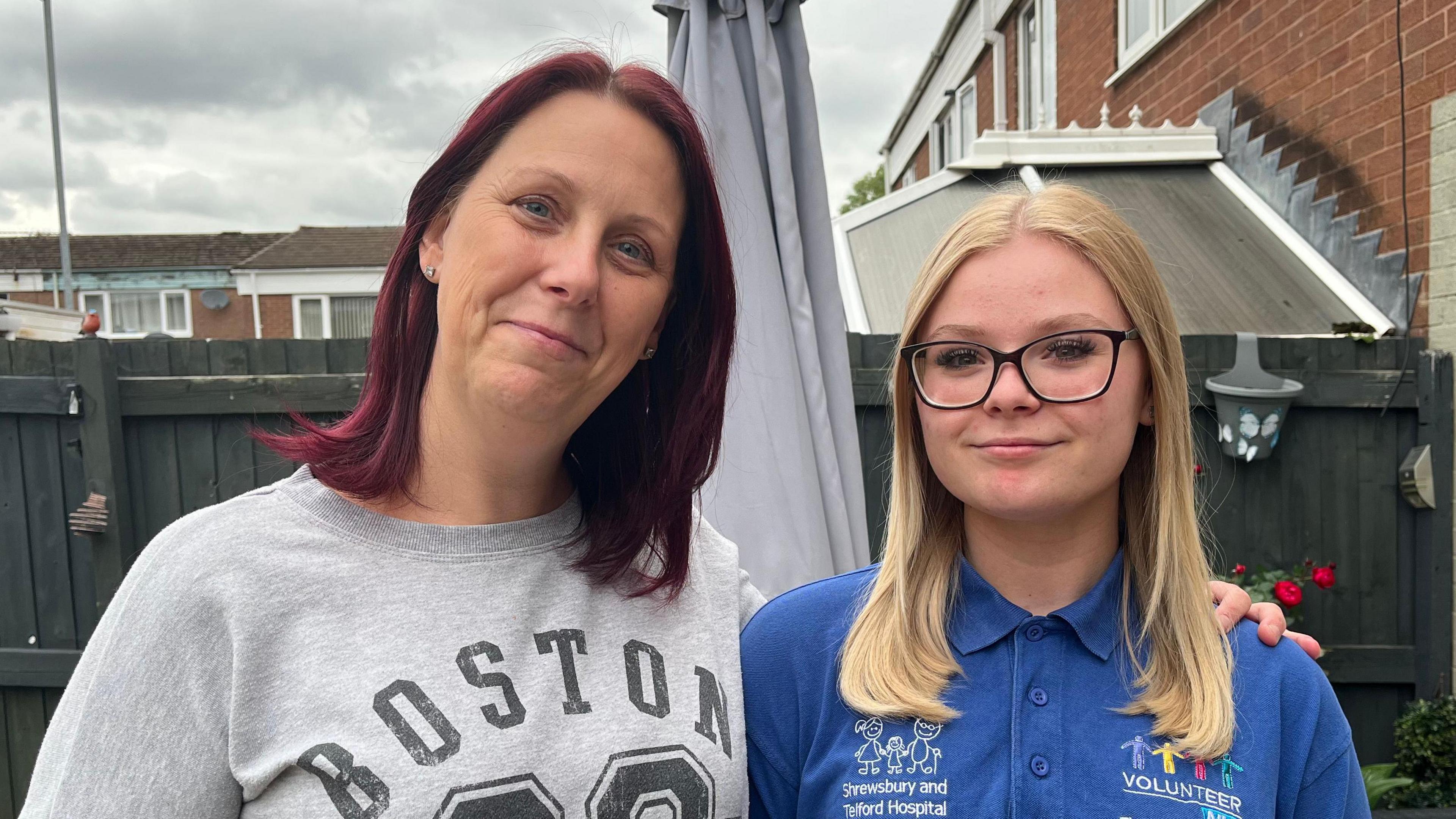 Sharon (left) and her daughter Alisha-Mai (right) stood side-by-side with their arms around each other. Sharon is wearing a grey sweatshirt with 'Boston' written on it in black letters, and Alisha-Mai is wearing a royal blue polo shirt with 'Shrewsbury and Telford Hospital' and 'Volunteer' written on it in white letters. They're stood in the garden of their home, with a dark green wooden fence behind them. The sky is grey and overcast. 