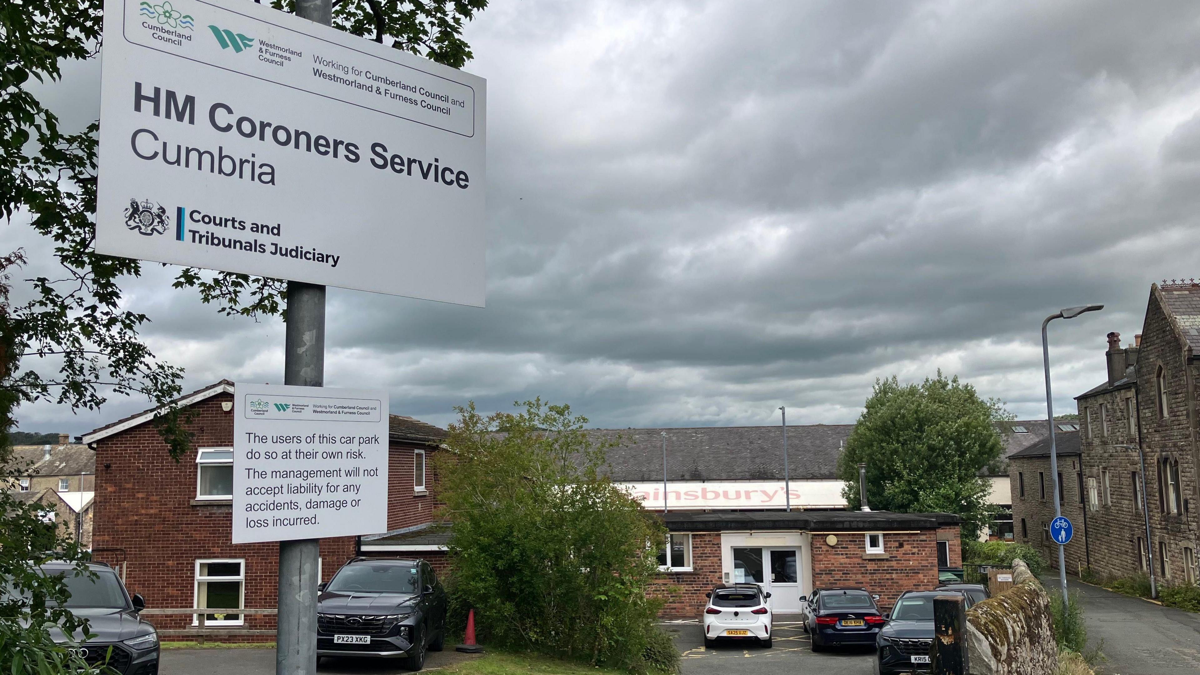 The sign at the entrance to the coroner's court in Cockermouth. It is a white sign on a silver metal pole with various logos and names of government organisations, with the most prominent words across the middle reading HM Coroners Service Cumbria. The gable end of a brick two-storey building with a pointed tiled roof stans at right angles to a single storey flat-roofed extension which appears to house the main entrance. Behind the smaller building is a Sainsbury's shop front. Cars are parked outside the coroners office. On the opposite side of a narrow street, with no pavement, to the right of the building building are much older two and three storey stone buildings in a terrace.