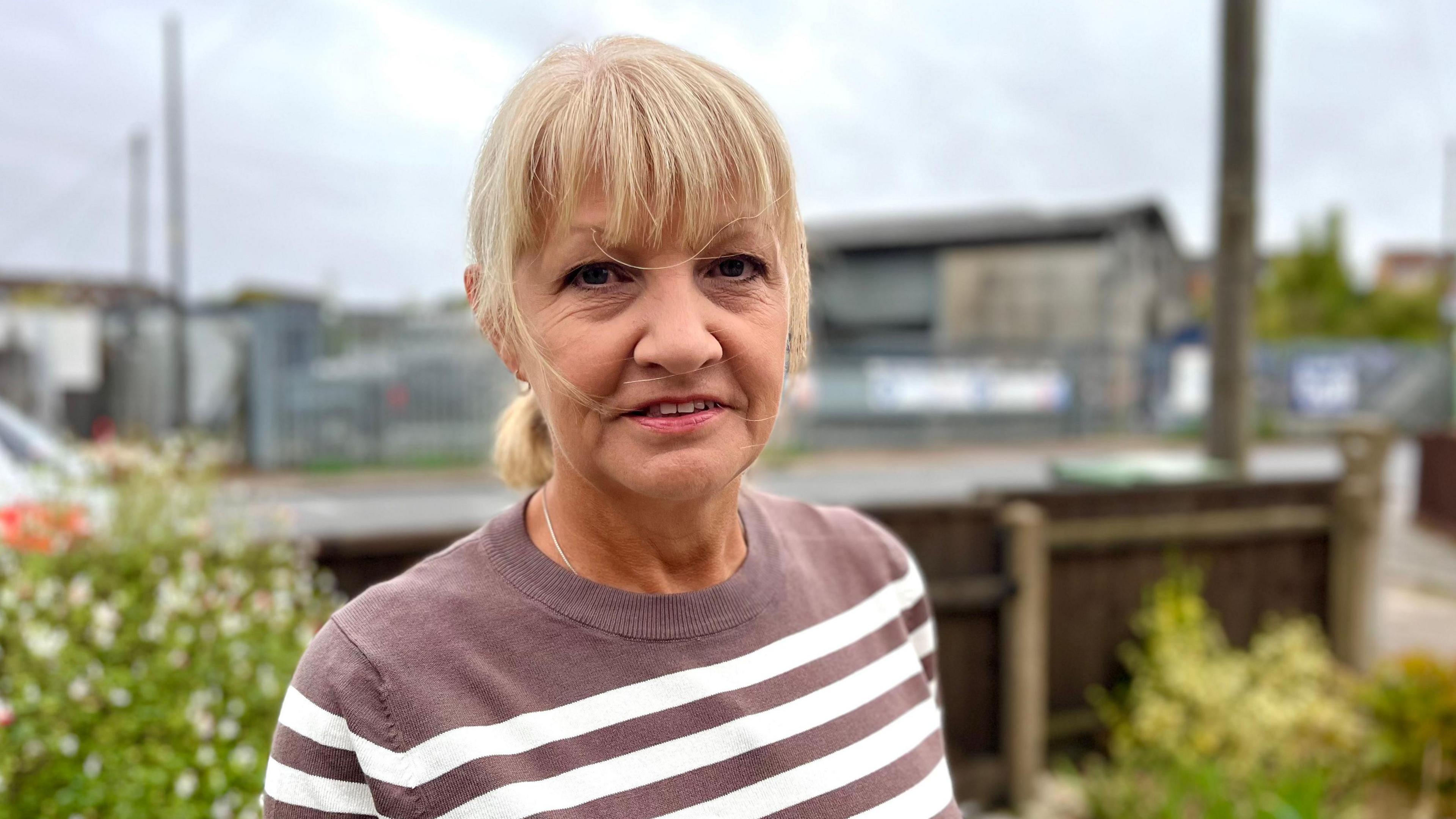 Sue Keeley with fair hair pulled back in a pony tail and a fringe, wearing a light brown jumper with white stripes and behind her, across a road, a warehouse