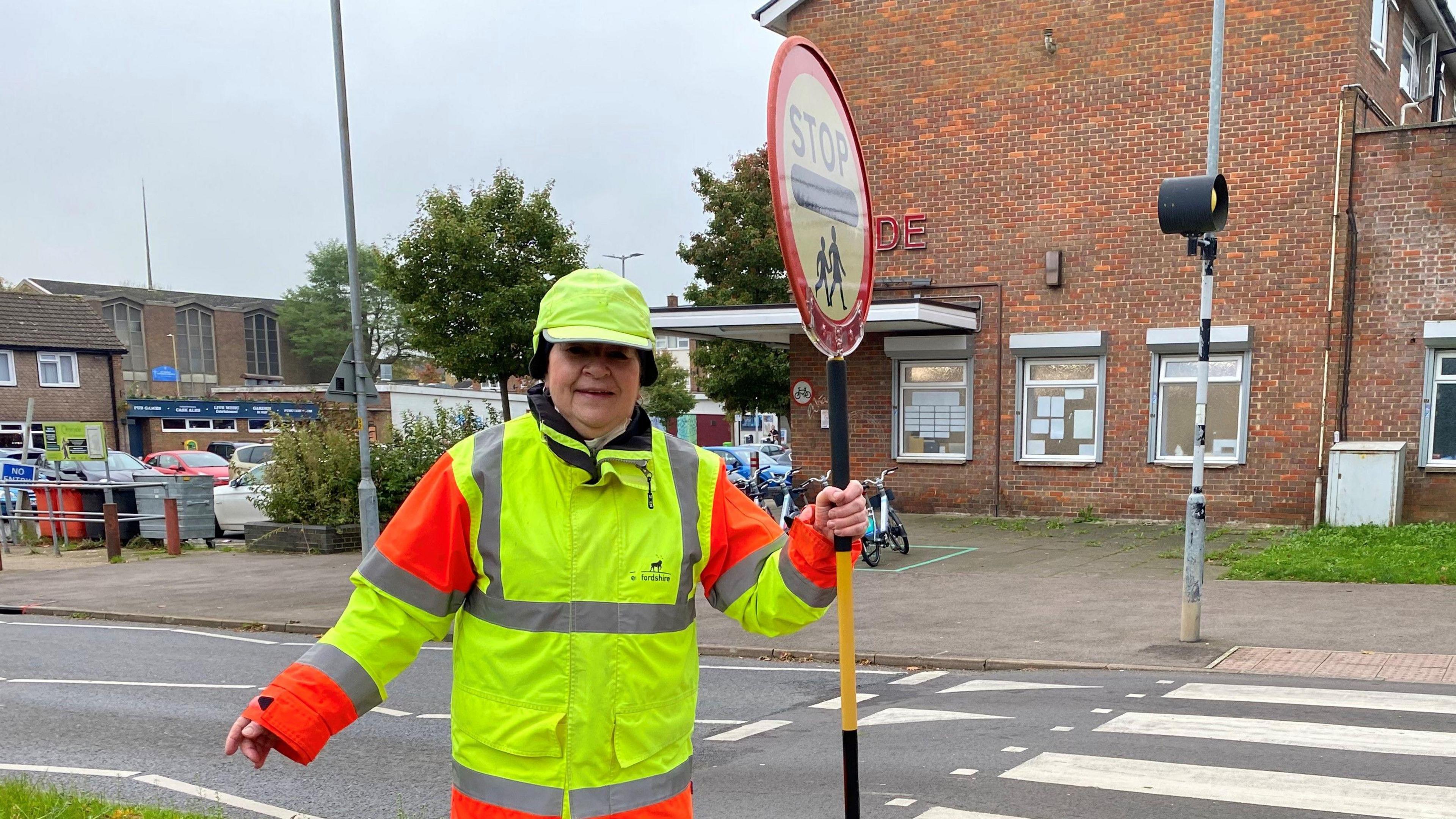 Julie Bennett, dressed as a lollipop lady, with a high vis hat, jacket and holding a lollipop patrol stick with STOP on it. She is standing by a road crossing with one arm to her side and the other holding her stick. She is looking straight at the camera.