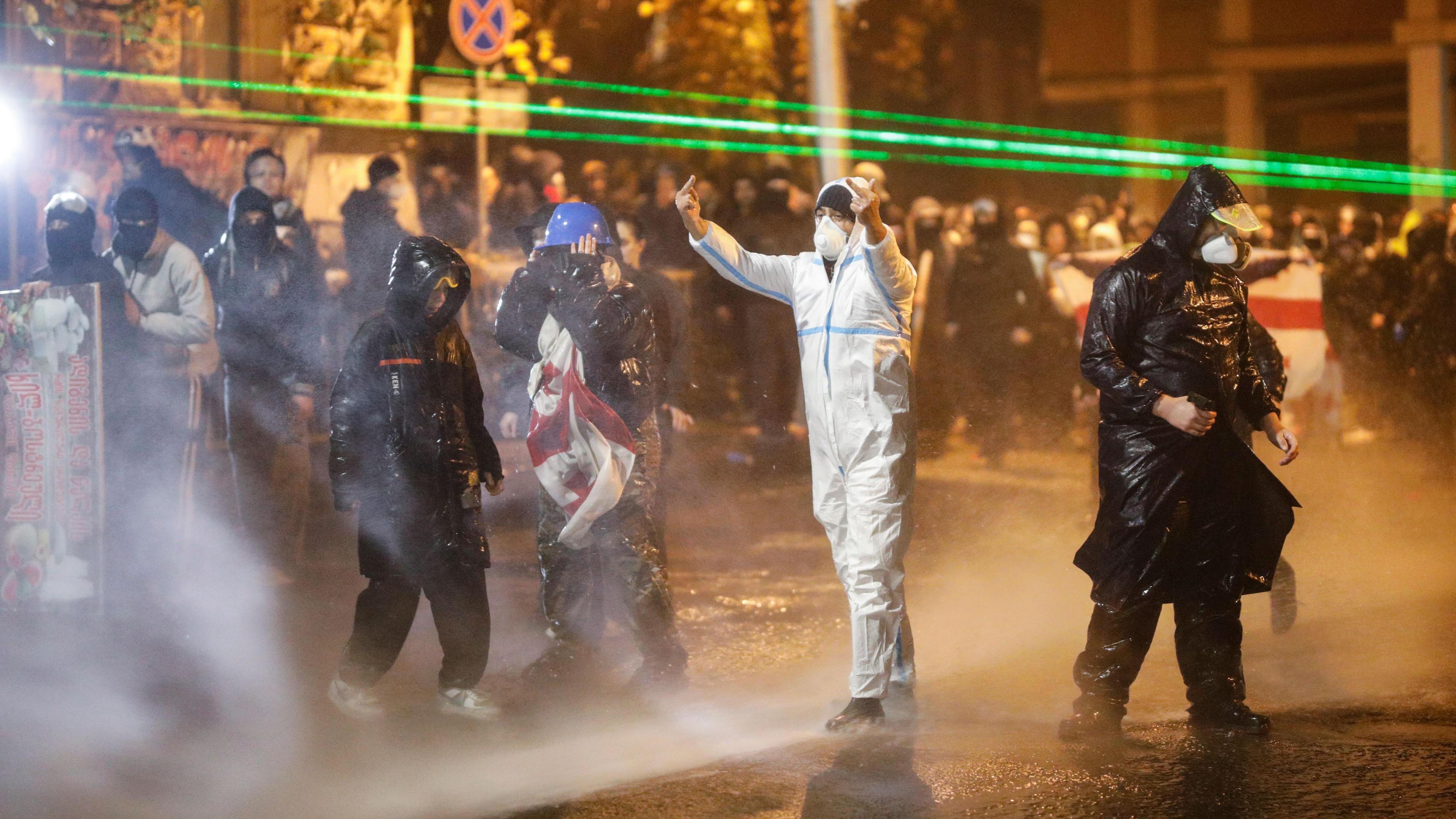 Protesters wearing rain jackets and hoods are pummelled with jets of water, as one of them swears at police with hand gesture.