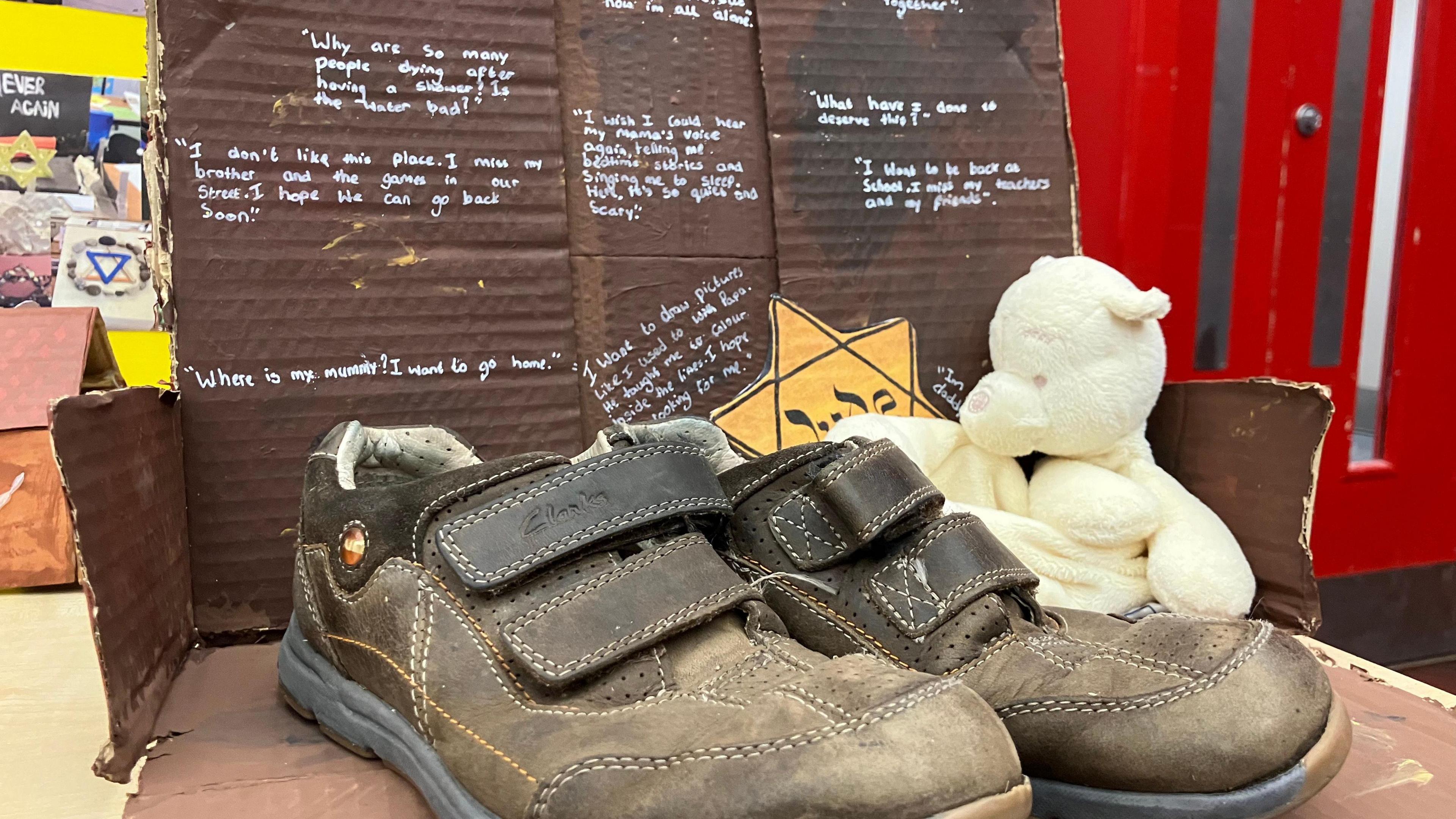 Some brown shoes, inside a brown painted cardboard box, with small text in silver writing, a teddy bear and a yellow star of david badge with Jude