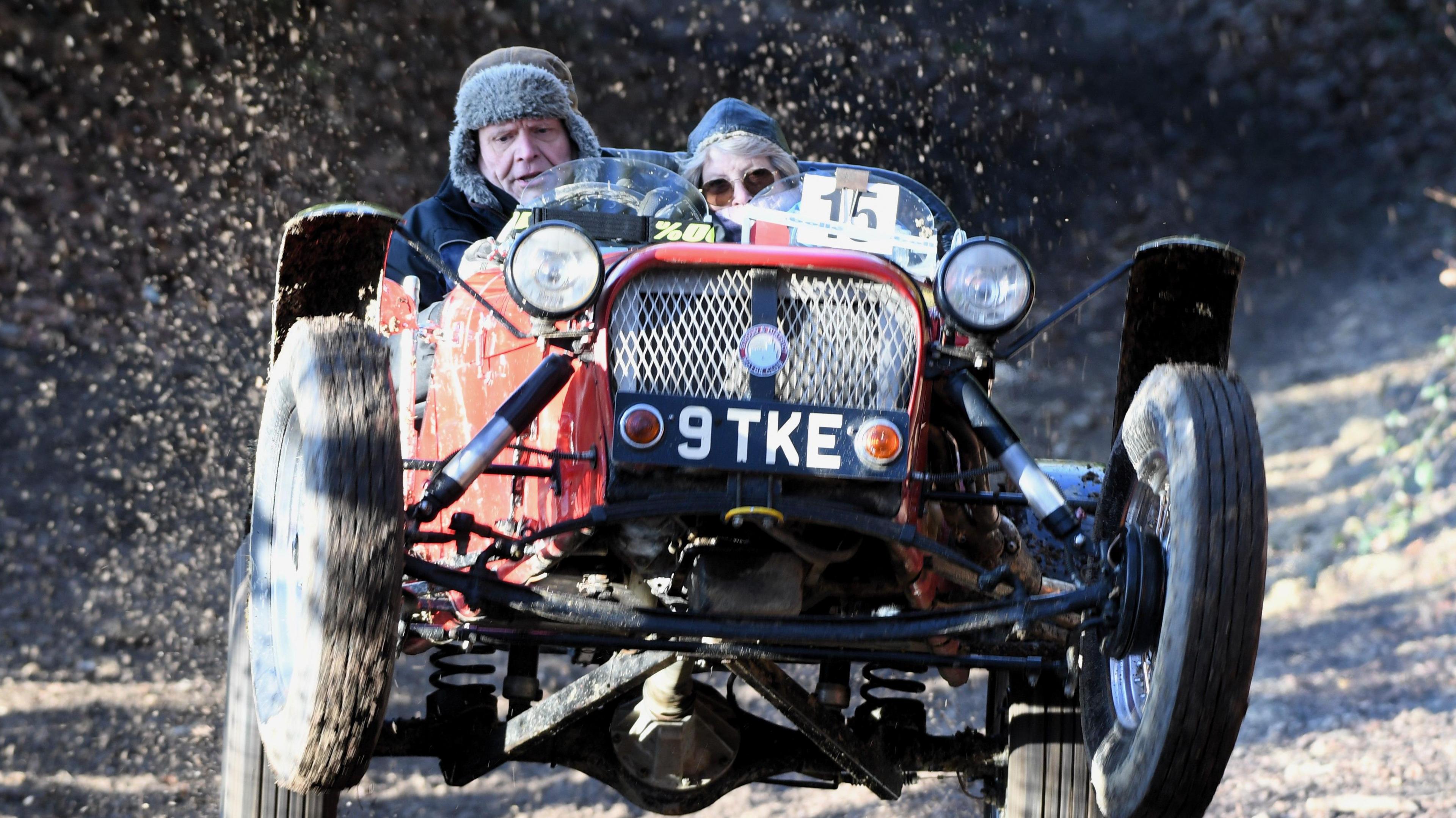 A open-top race car lurches forward with its front wheels in the air. The driver and a passenger, both wearing hats, are huddled behind a low windscreen as mud flies up.