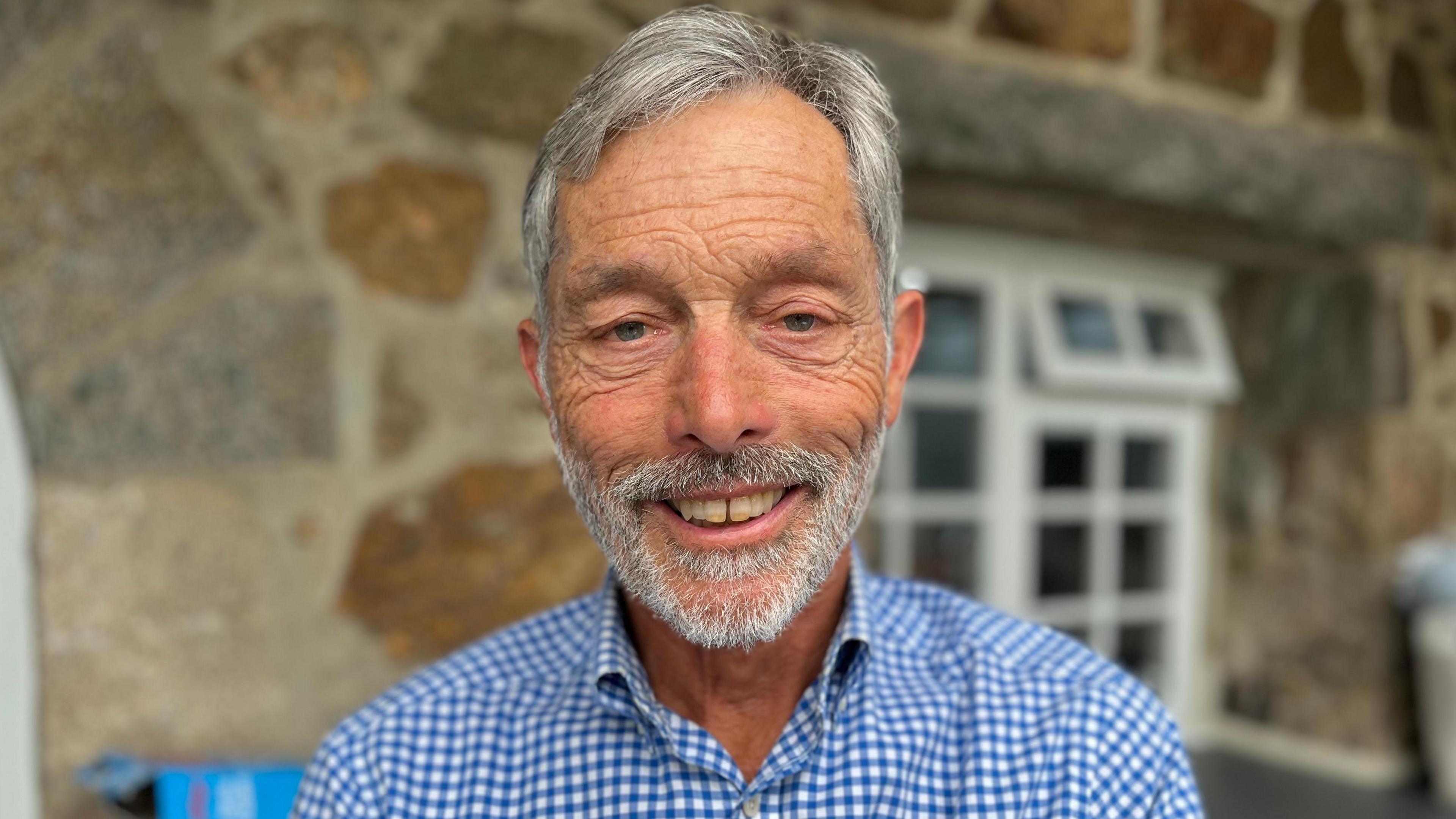 Andrew Ozanne, with a grey beard smiling towards camera wearing a blue and white checked shirt.