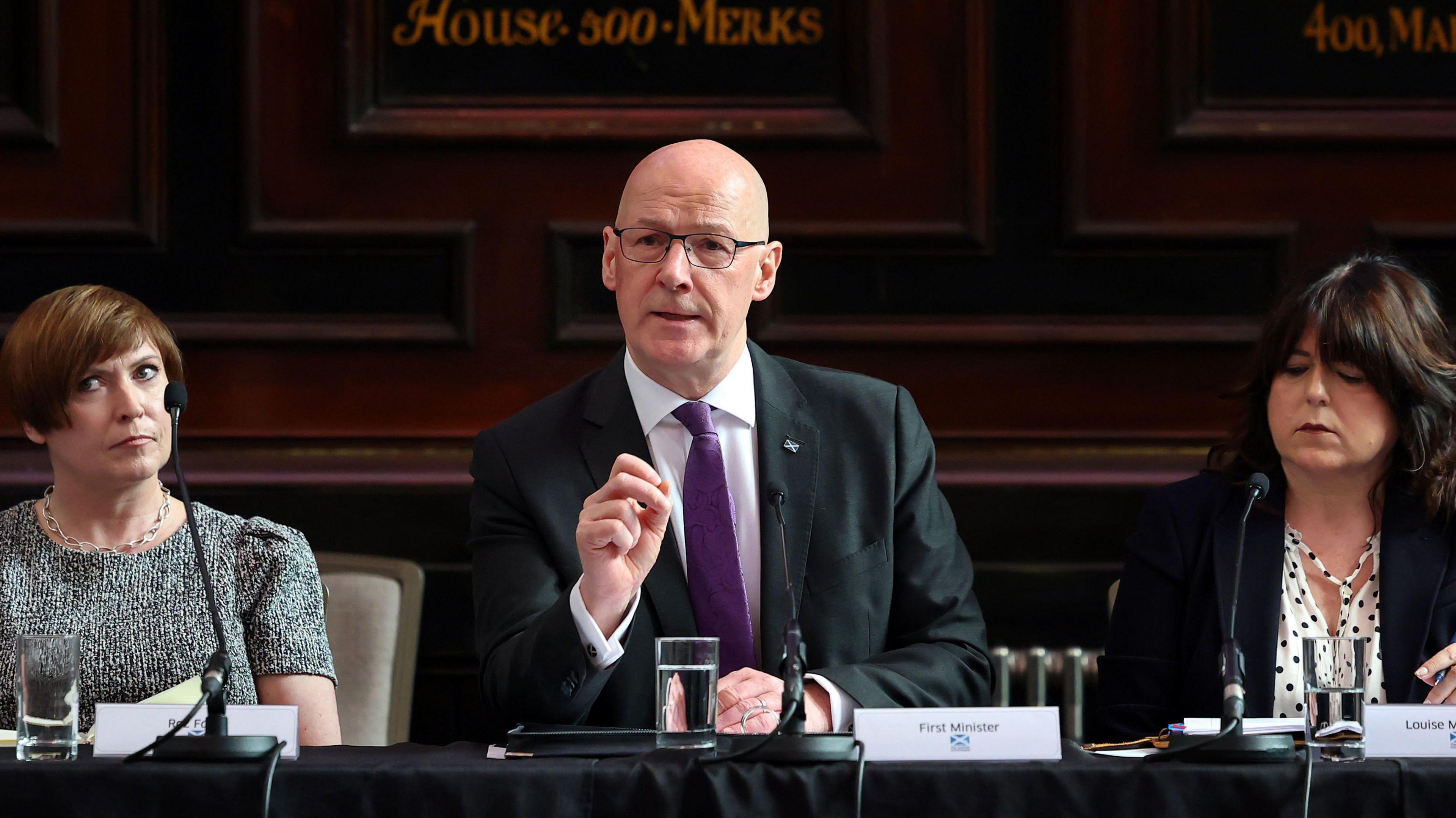 John Swinney - wearing a dark suit with purple tie - sits behind a desk, with a microphone and glass of water in front of him. He has his hand raised. Two women sit next to him listening - one is wearing a grey top and the other with a white top and black jacket.