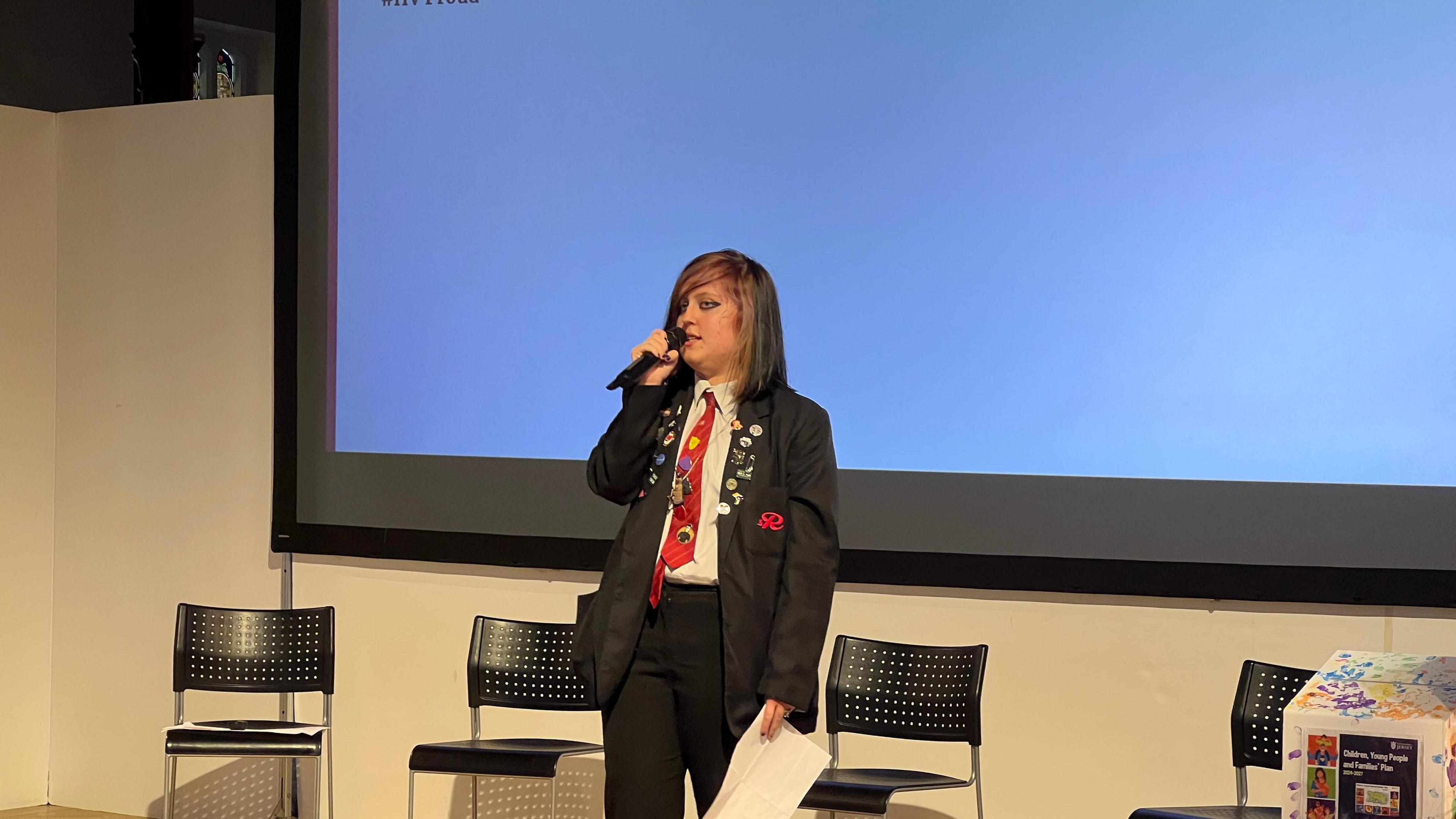 Lexi stands on a stage with a sheet of paper in one hand and a microphone in the other. Behind her are four black chairs and there's a white projection screen on the wall.