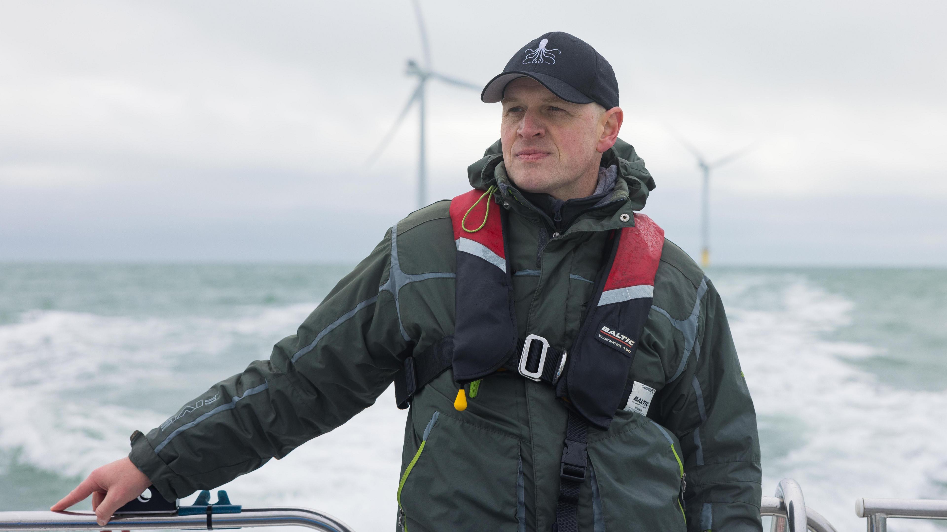 A man standing on a boat out a sea. The boat is not visible. The man is facing the camera but looking off to his right, our left. His right hand is on a metal railing and his left hand is by his side. He is wearing a dark green jacket, a black and red left jacket, and a black cap. The sky behind him is grey, the sea is grey and white from the wash of the boat, and there are two large wind turbines behind him also