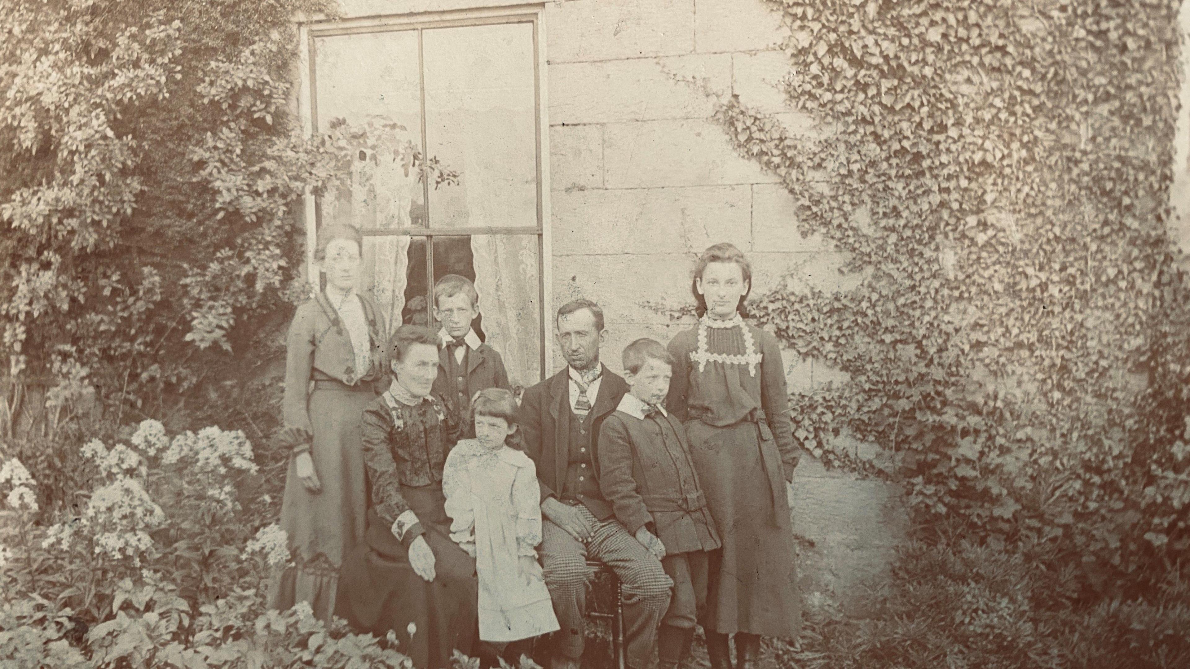 A black and white picture of a family group sitting and standing in front of a window. It looks to date from the very early twentieth century.