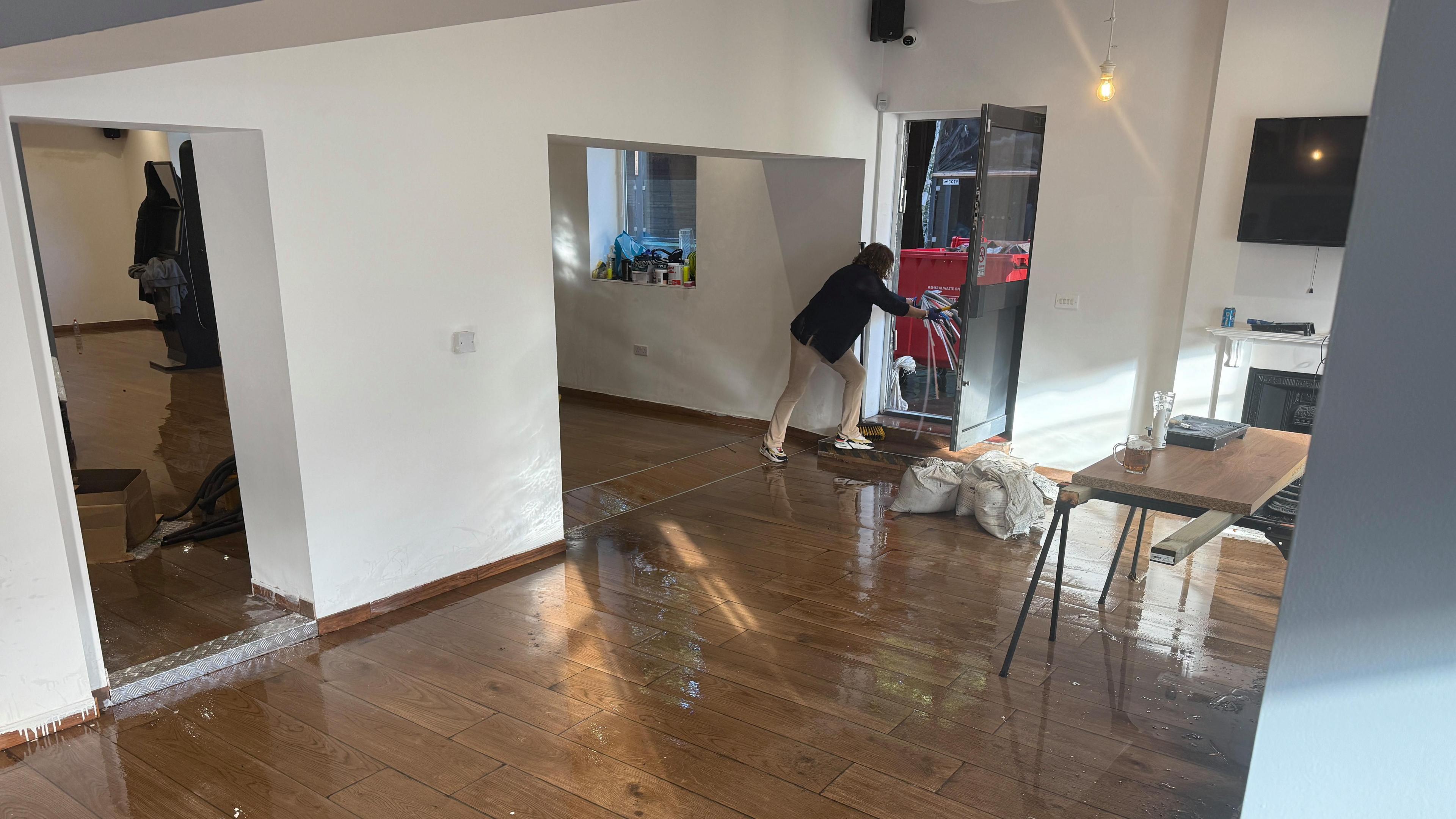 woman disposes of rubbish from inside a flooded pub. There are sandbagson the floor and the wooden floor is soaked with water.