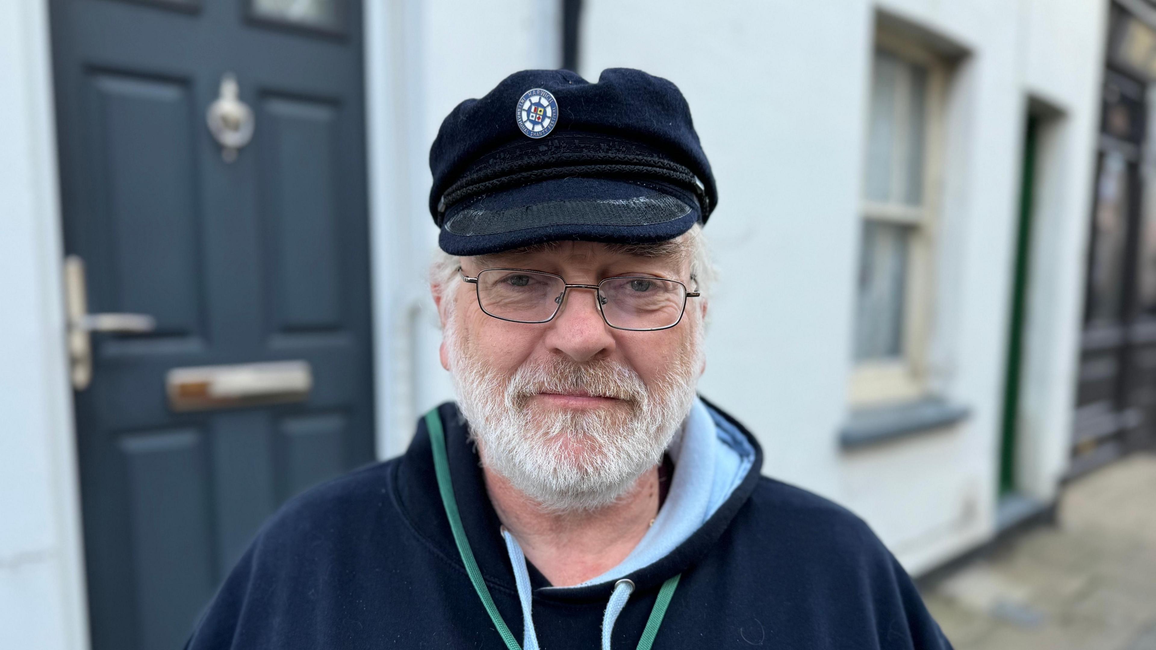 A man looking straight at the camera. He is wearing a navy hoodie with a green lanyard around his neck and a navy hat on his head.