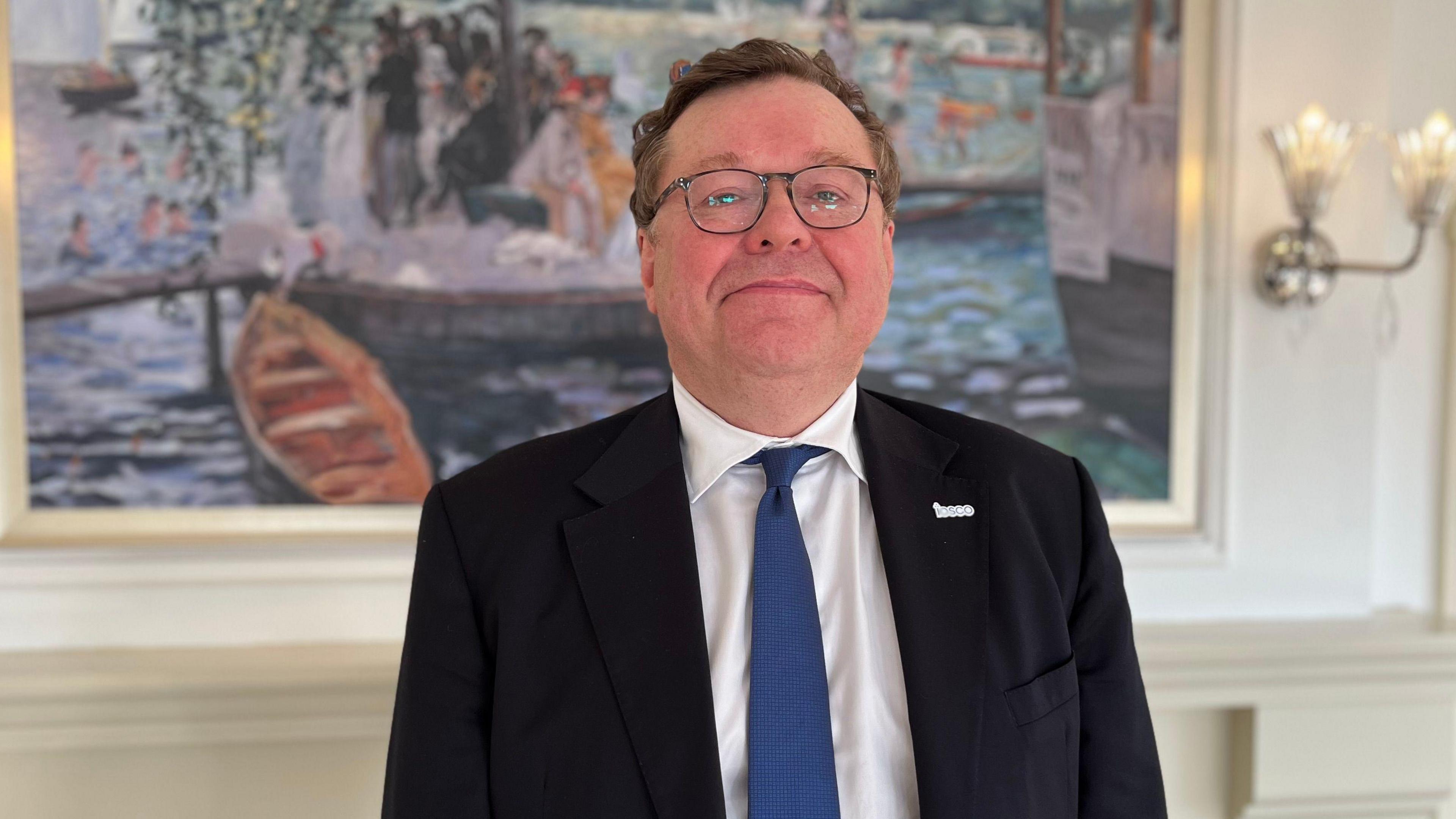 A white man with a black suit and a blue tie wearing a white shit and a pin with the letters IOSCO on his left lapel. He is smiling and wearing glasses. 