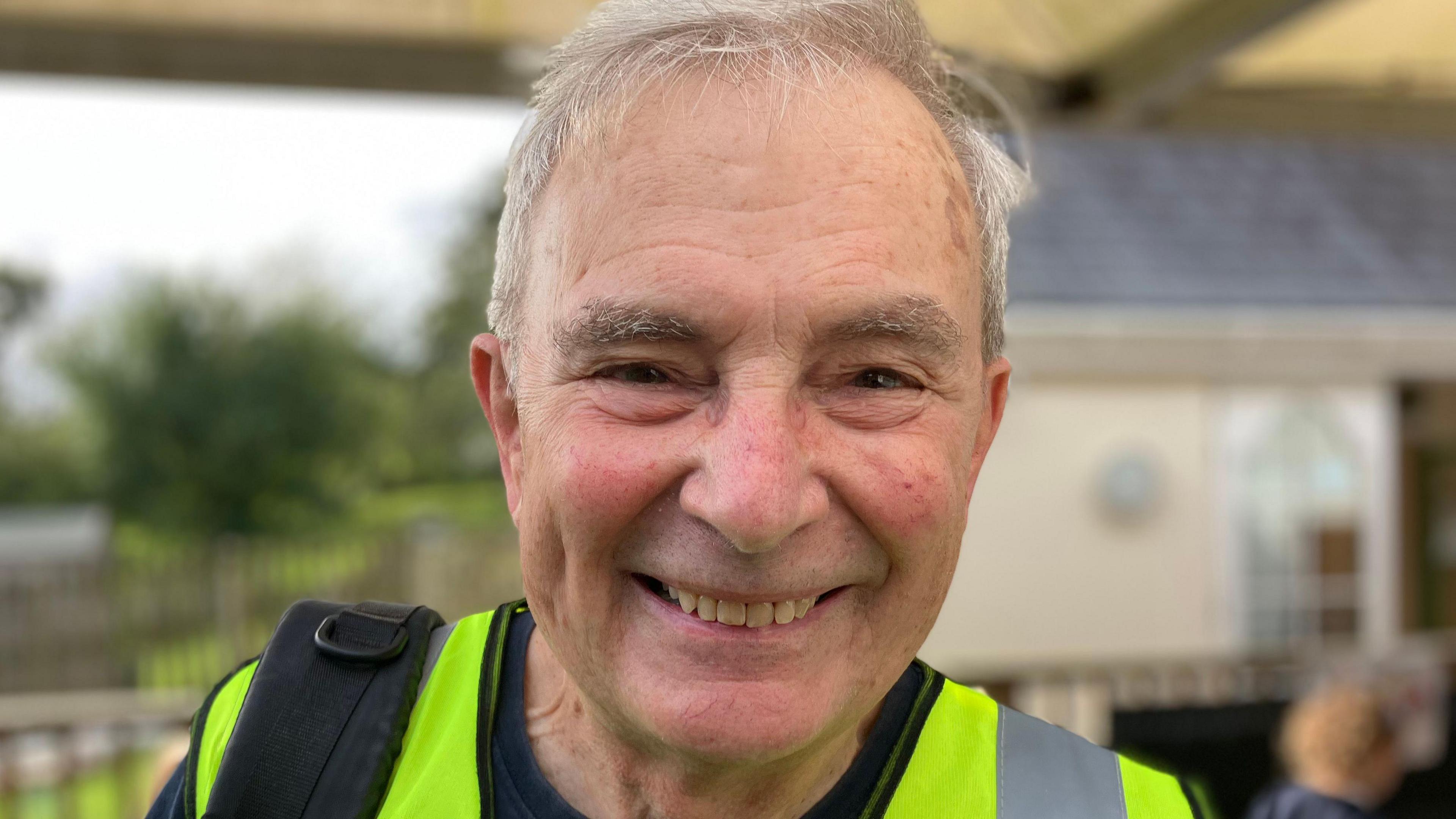 An older man in a high visibility yellow jacket smiles.