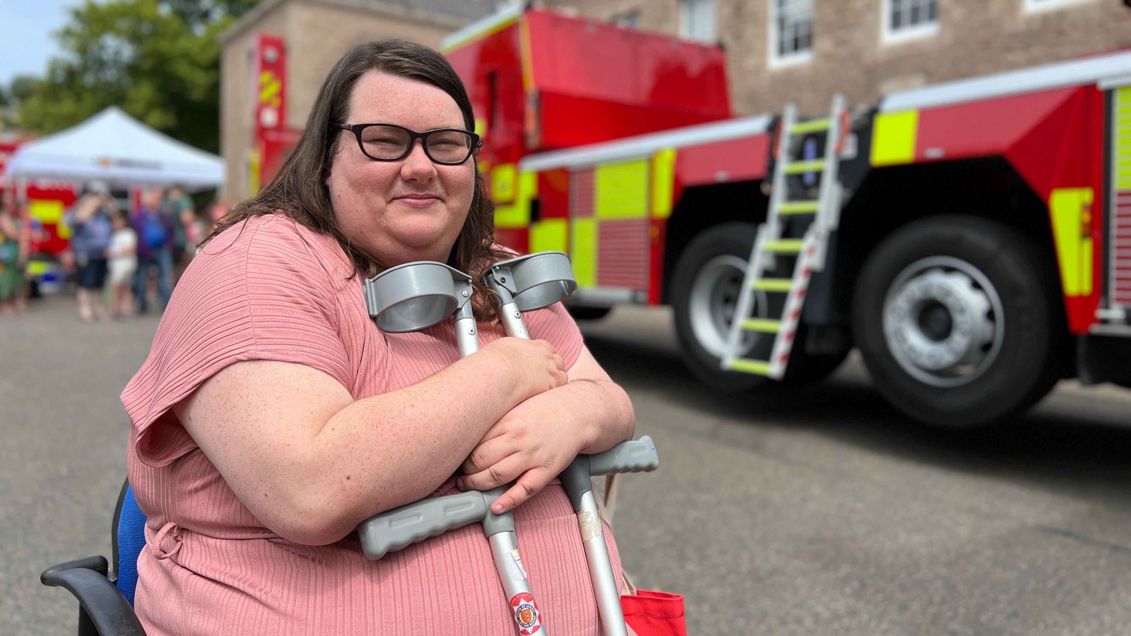 Vicky smiles at the camera as she sits in a chair holding her crutches. Behind he is a fire engine and crowds looking at stalls. She is wearing a pink dress.
