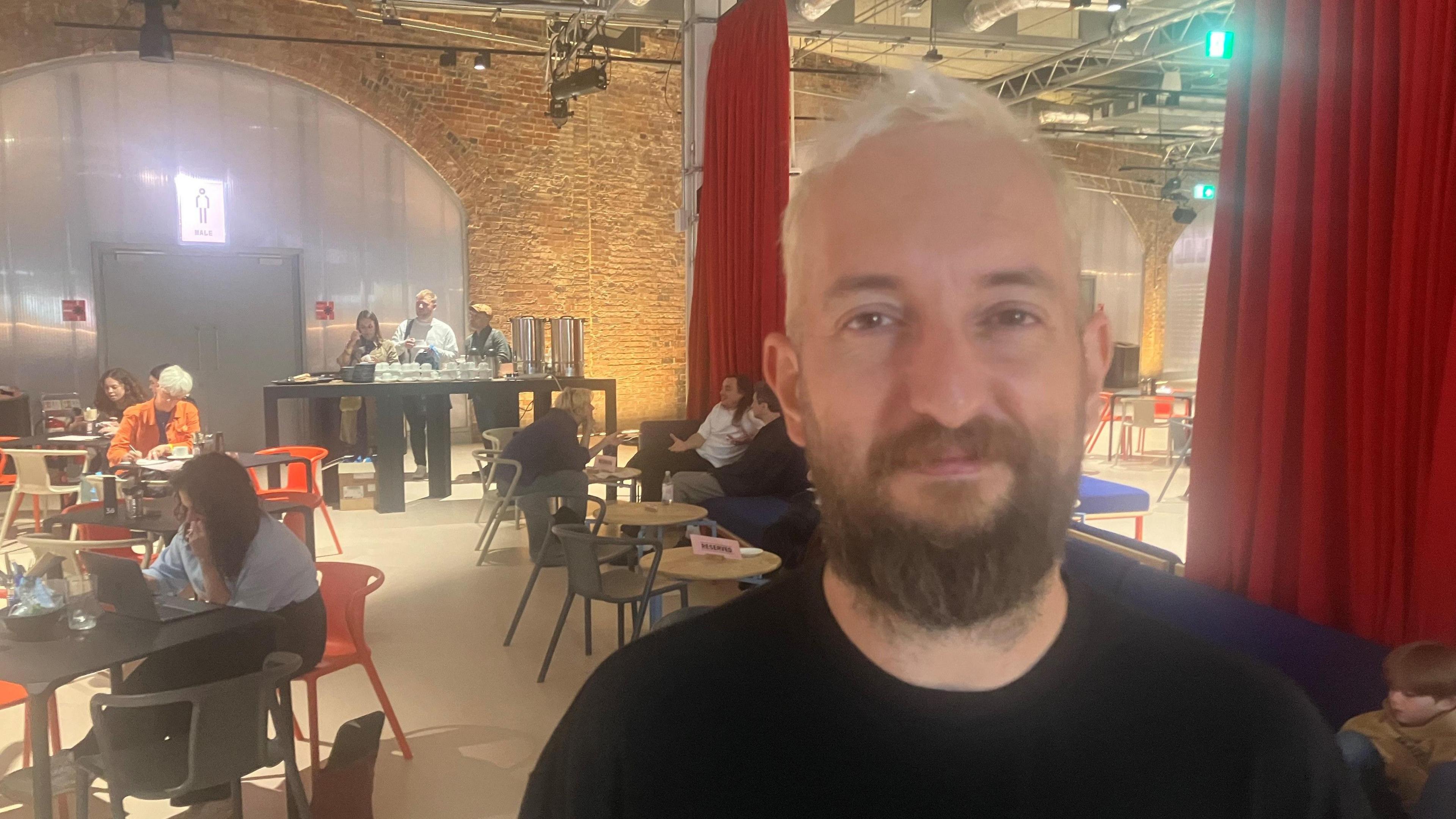 Matthew Bessudo at the Aviva Studios venue, standing in a communal area with people working on laptops at tables behind him