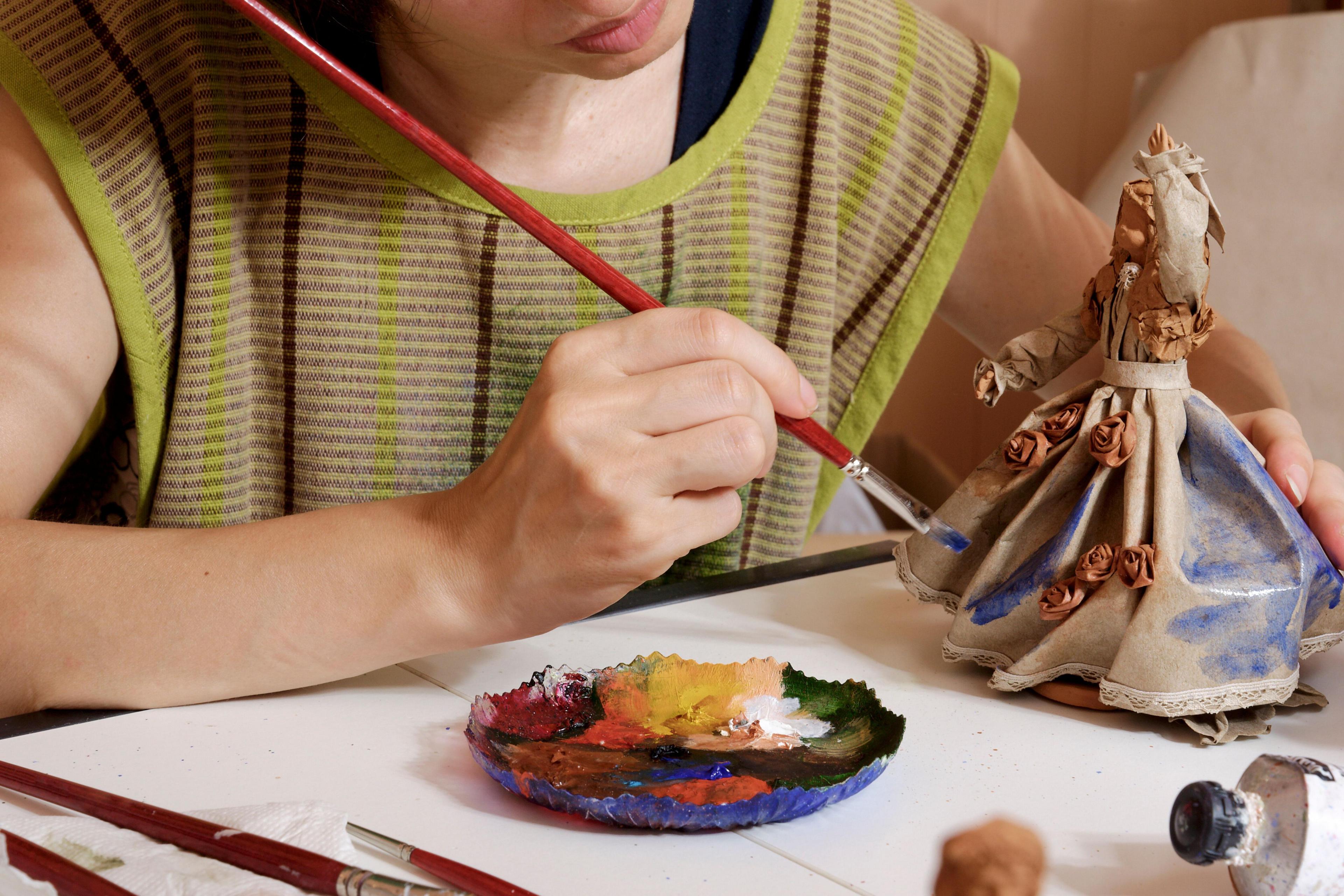 An artist paints a figurine. She is holding a paintbrush in one hand and has a dish with paint in front of her on a covered table. The dish has different colours in it, including reds, yellows and white. The ceramic figurine is partly painted with her hand raised and has a long flowing dress.