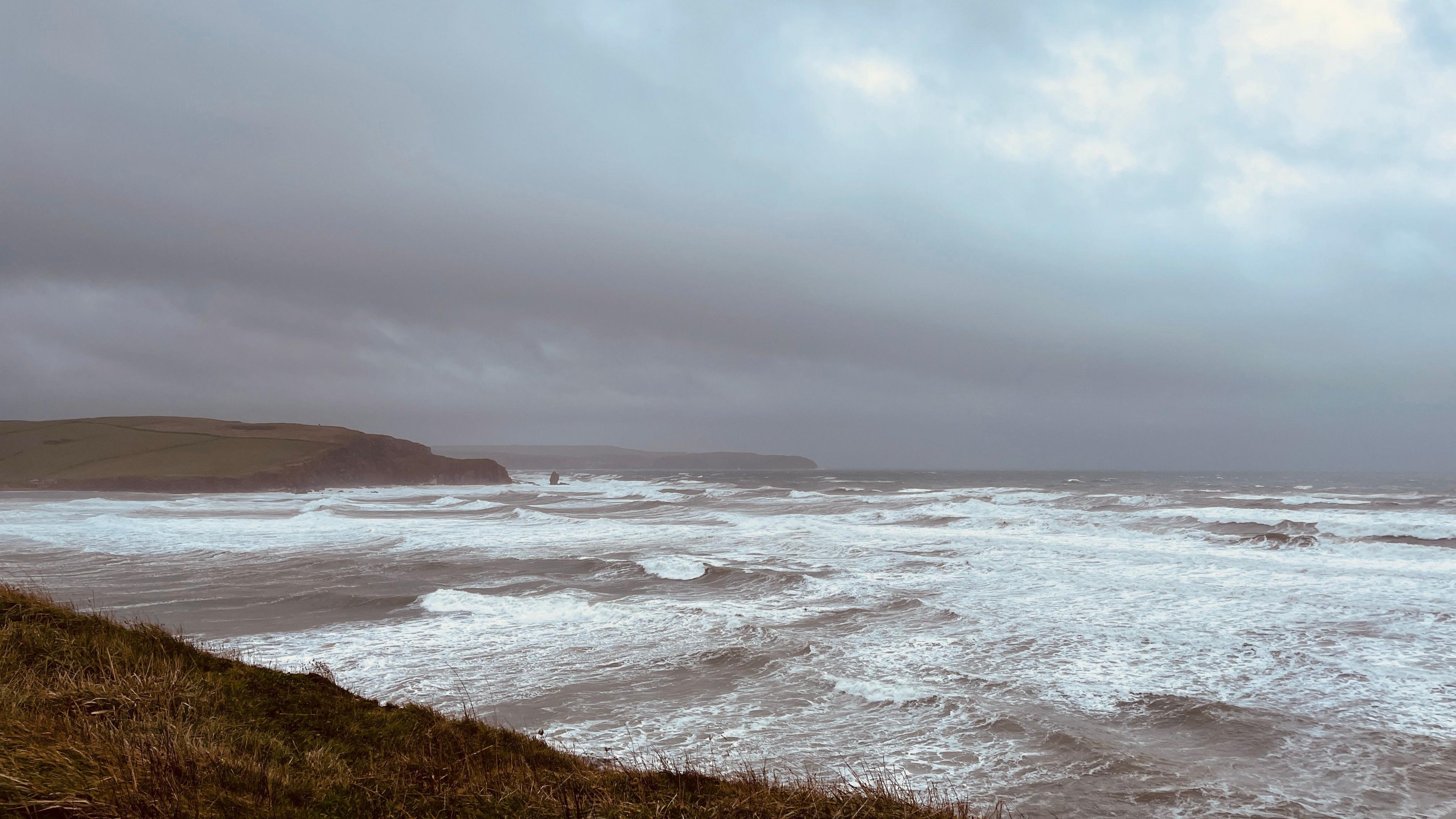 A stormy sea over a wide beach in front of a grassy headland.