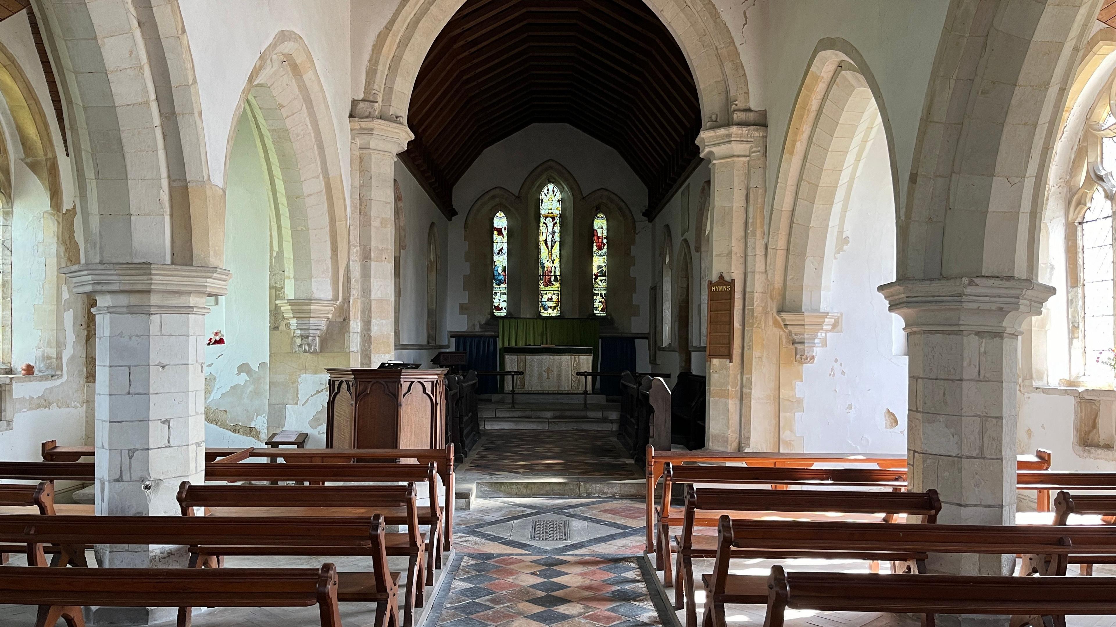 The inside of a church shows wooden pews and a tiled floor. There are stone arches to the sides and a stained glass window at the end