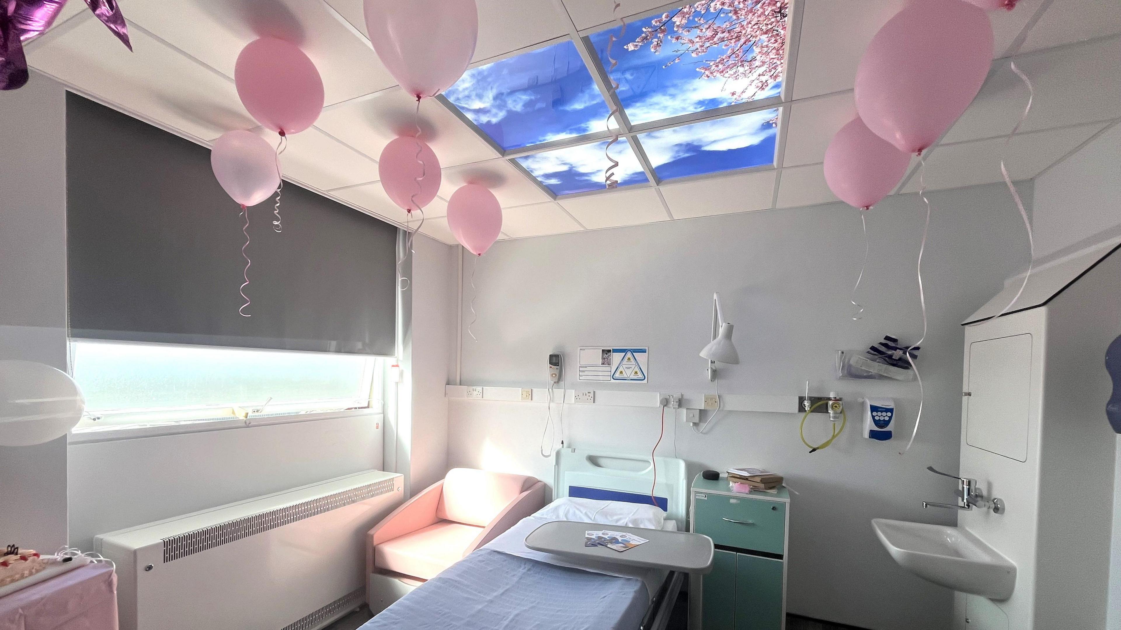 The "blossom room", which looks like a hospital room with some cherry blossom coloured furniture and a screen in the ceiling that looks like a sky window, looking up at a cherry blossom tree. There are pink balloons in the room too.