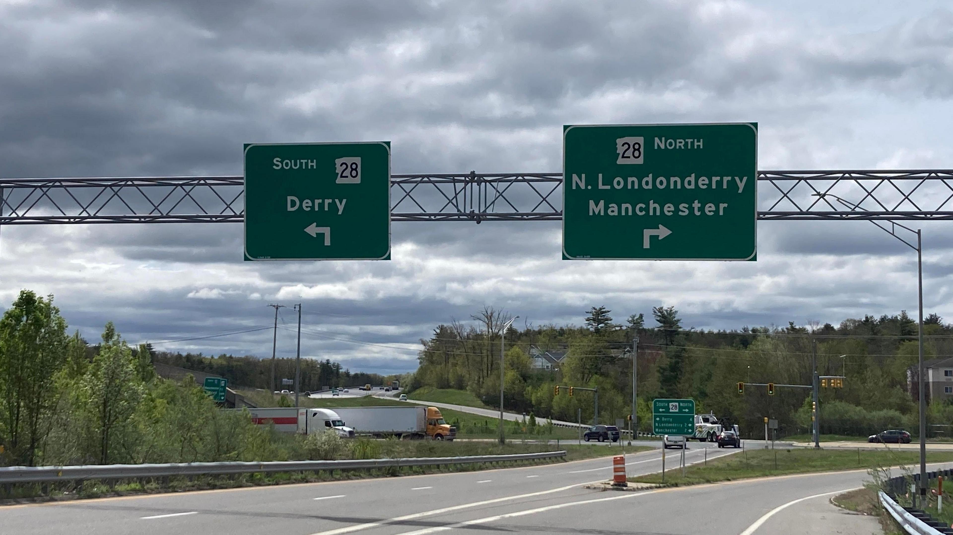 Two signs above a road in New Hampshire, one pointing left says south and Derry, the other pointing to the right says north N Londonderry and Manchester. There is a forest beside the road 