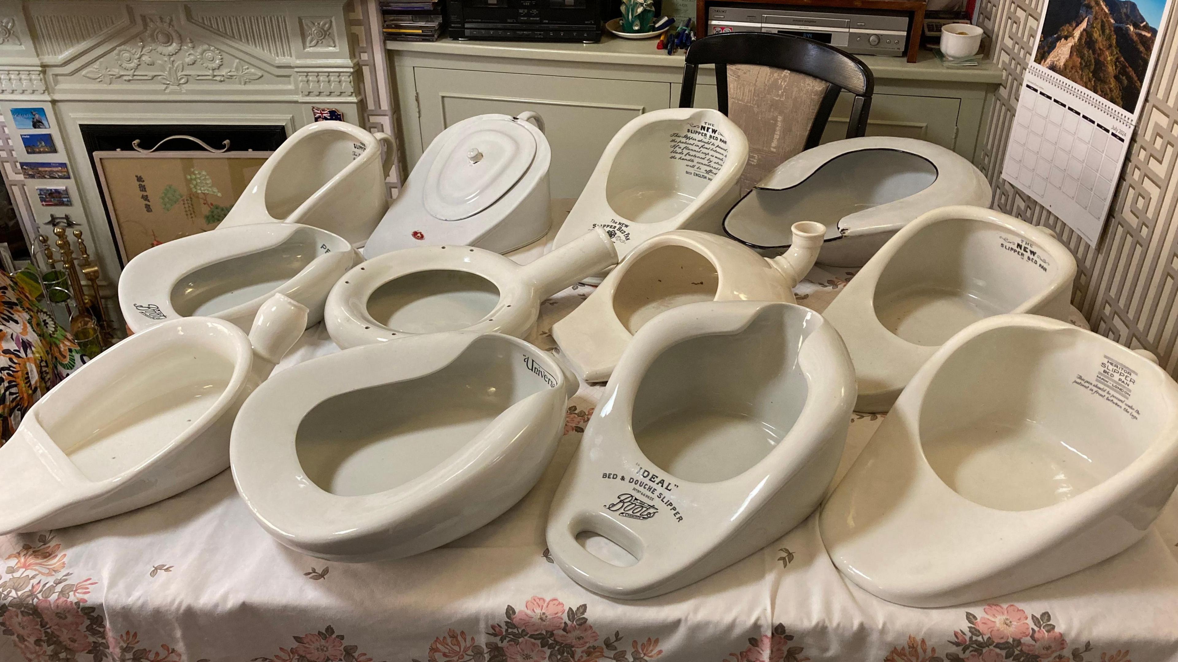 A closer view of the bedpans sitting on a kitchen table. They are all white and in different shapes and sizes. Some have writing and company logos printed upon them. 