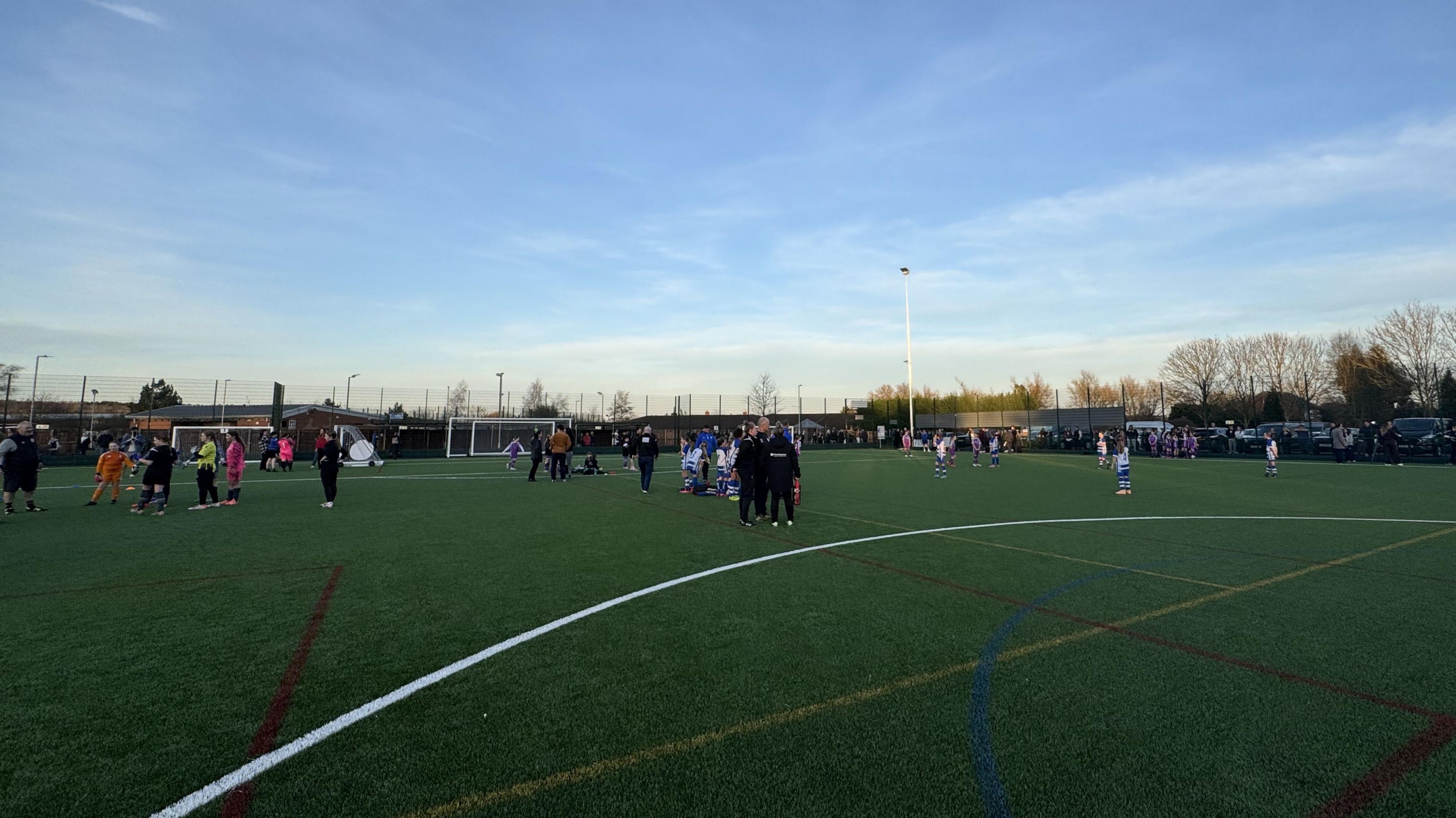 A wide image of the new Mary Earps Pitch in Calverton under a blue sky. The artificial grass is green and has yellow, blue, red and white lines marked on it. There are lots of people standing on it.