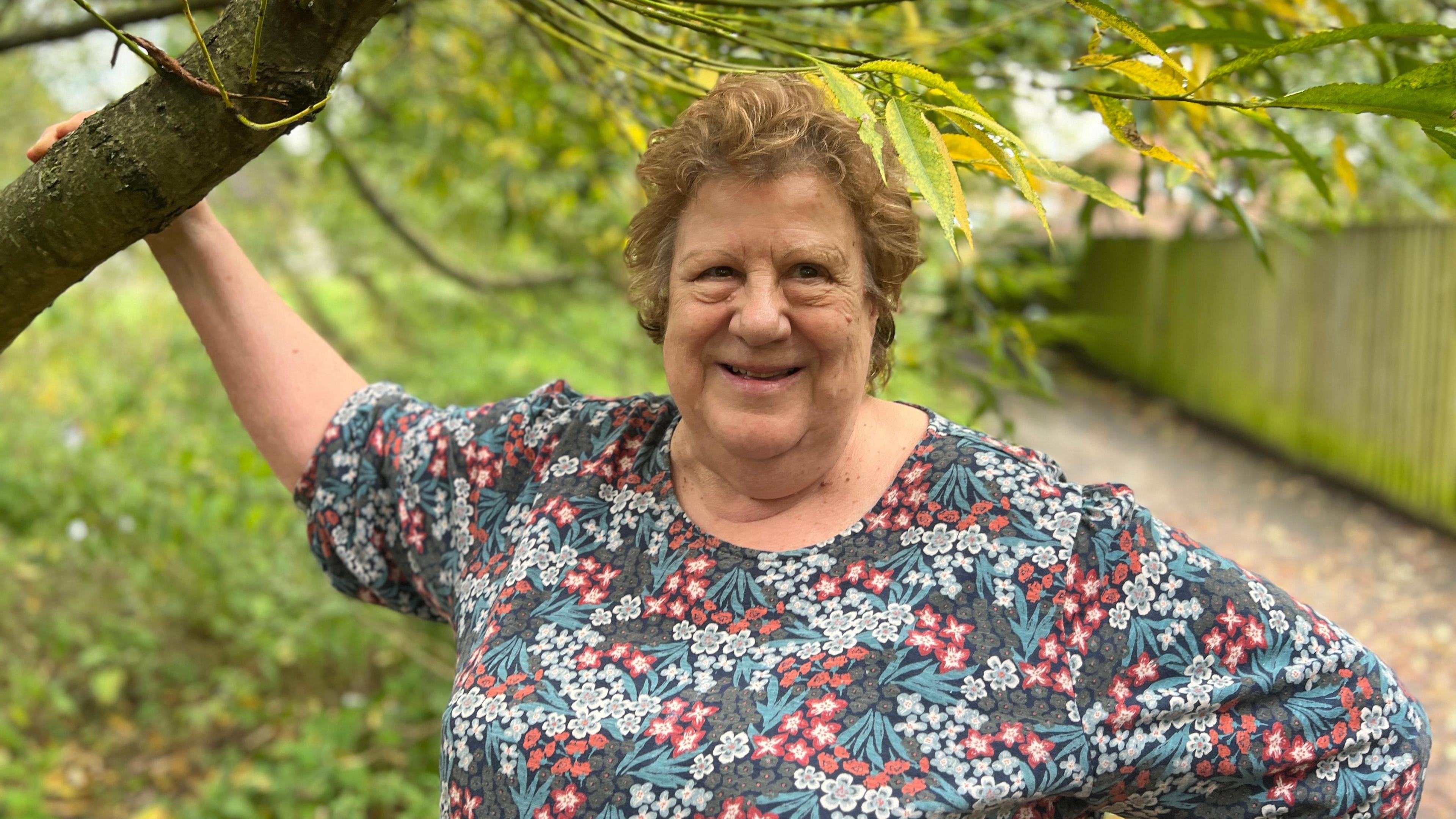 A lady with short curly blonde hair wearing a flowery blouse stands in a wooded area with one hand holding onto a tree branch above her.