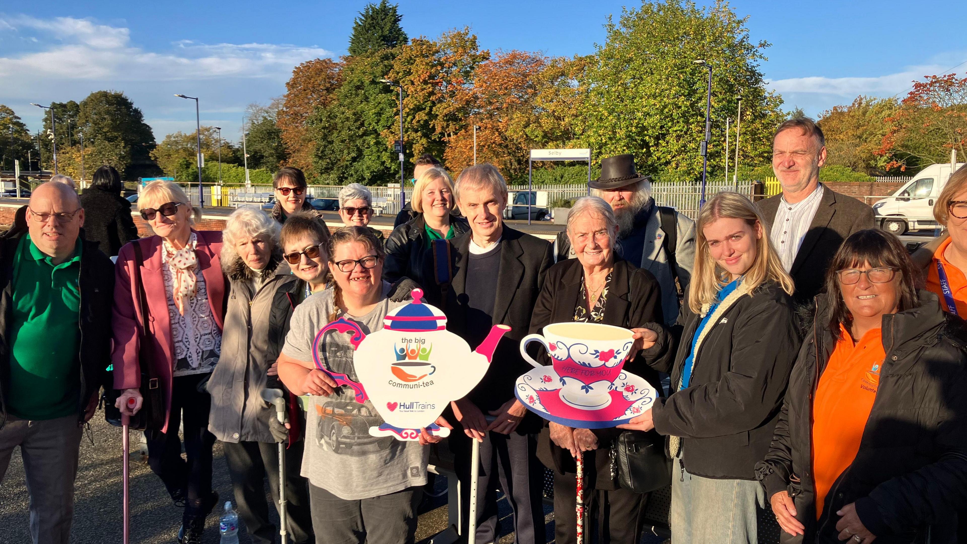 Staff and clients from mental health charity The Big Communi-Tea are waiting on the platform at Selby Station. They are holding a giant cardboard teapot and tea cup.