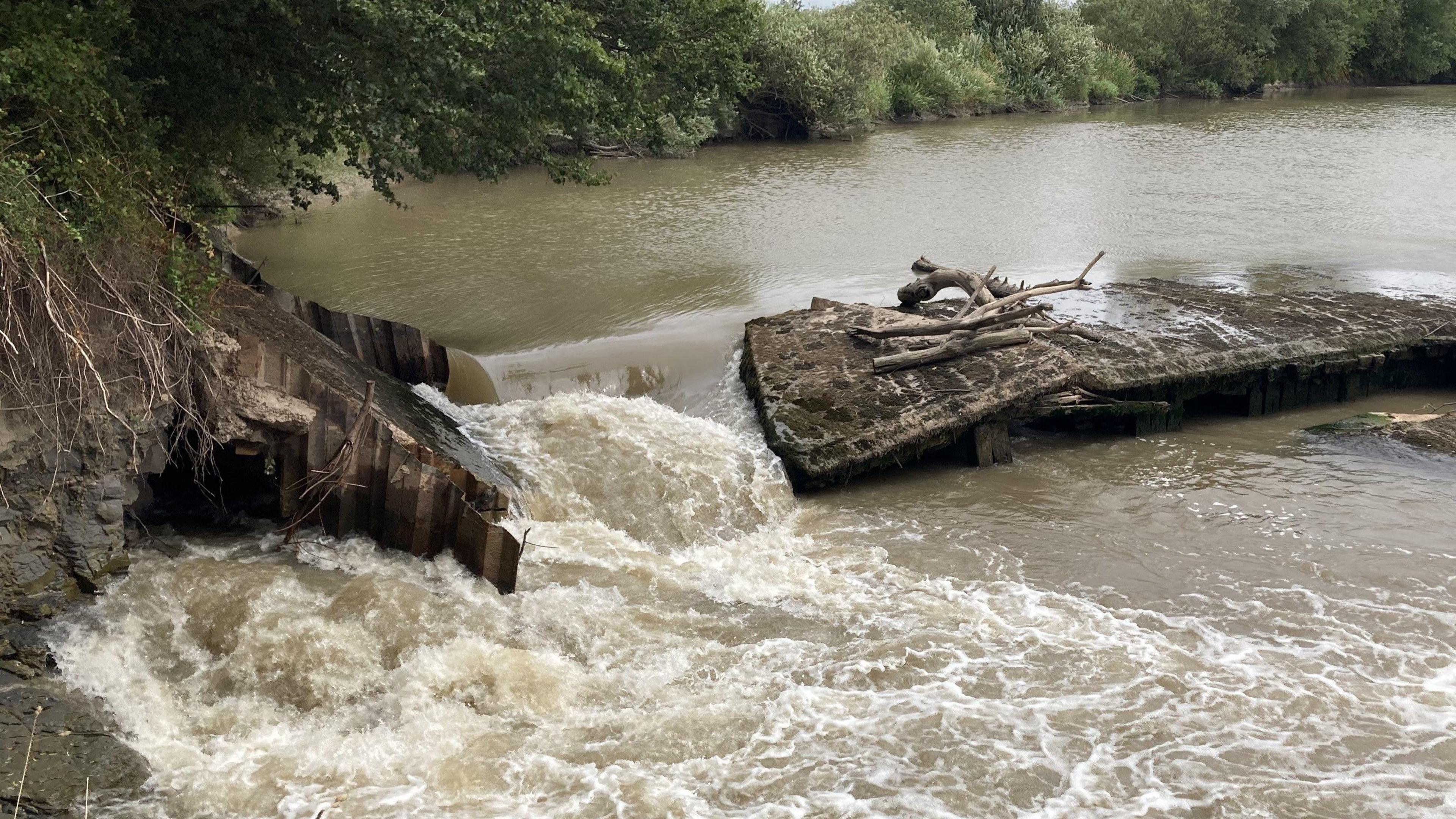 A photo showing a concrete weir across the River Severn. There is a large hole to the left side of the weir with water pouring through
